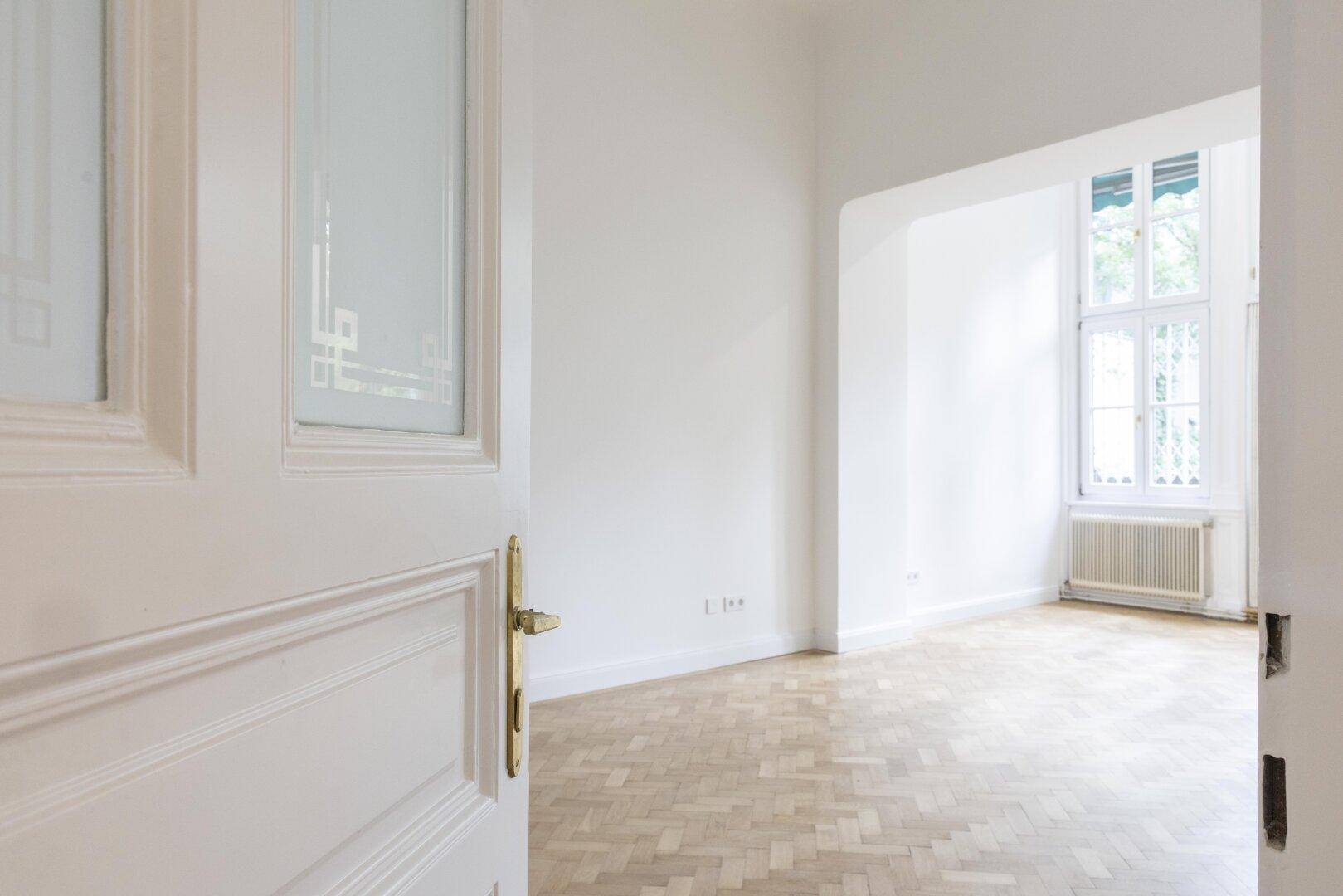 Helles, leeres Zimmer mit Fischgrätparkett und einem großen Fenster, das viel Tageslicht hereinlässt.