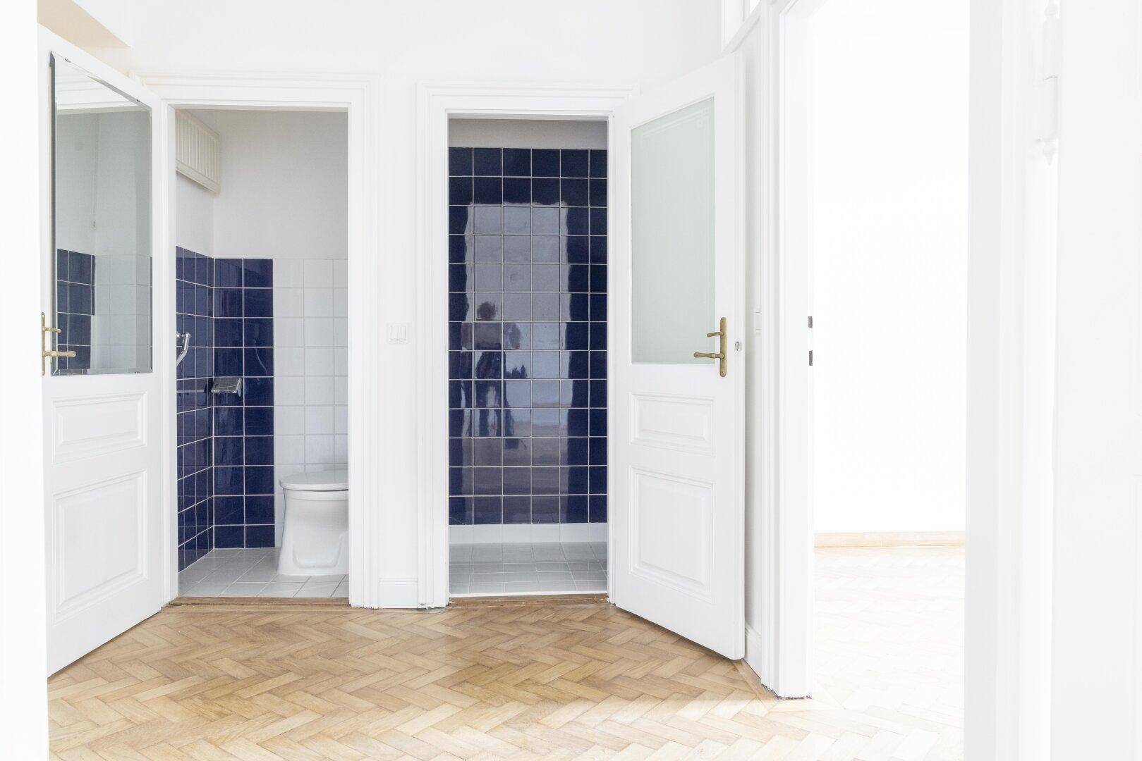 Zwei separate, geflieste Bereiche mit dunklen Fliesen, einer mit Toilette und der andere mit Dusche.