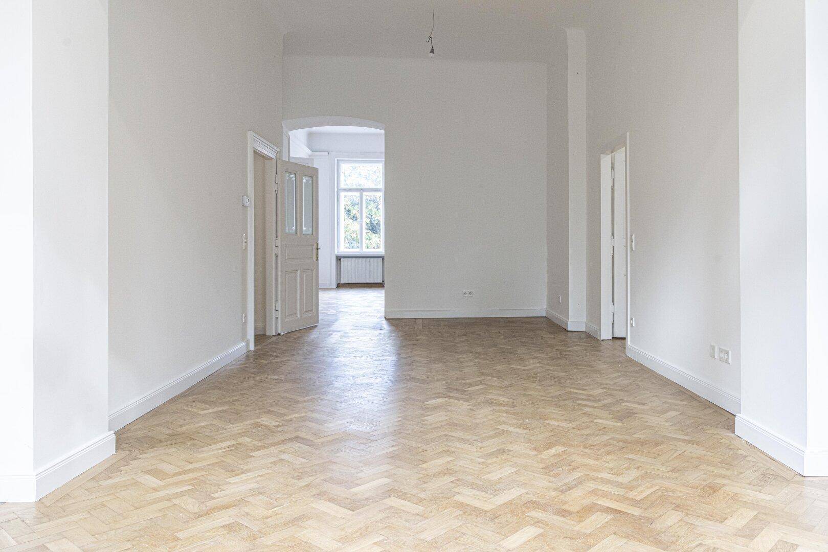 Heller Raum mit Fischgrätparkett und einem eleganten Torbogen, der zu einem weiteren Zimmer führt.