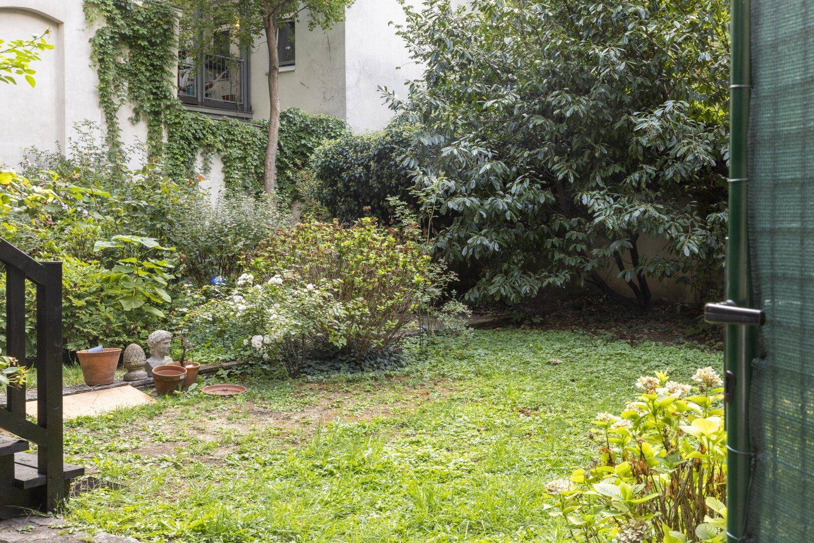 Gepflegter Gartenbereich mit Rasenfläche, verschiedenen Pflanzen und dekorativen Töpfen, umgeben von Grün.