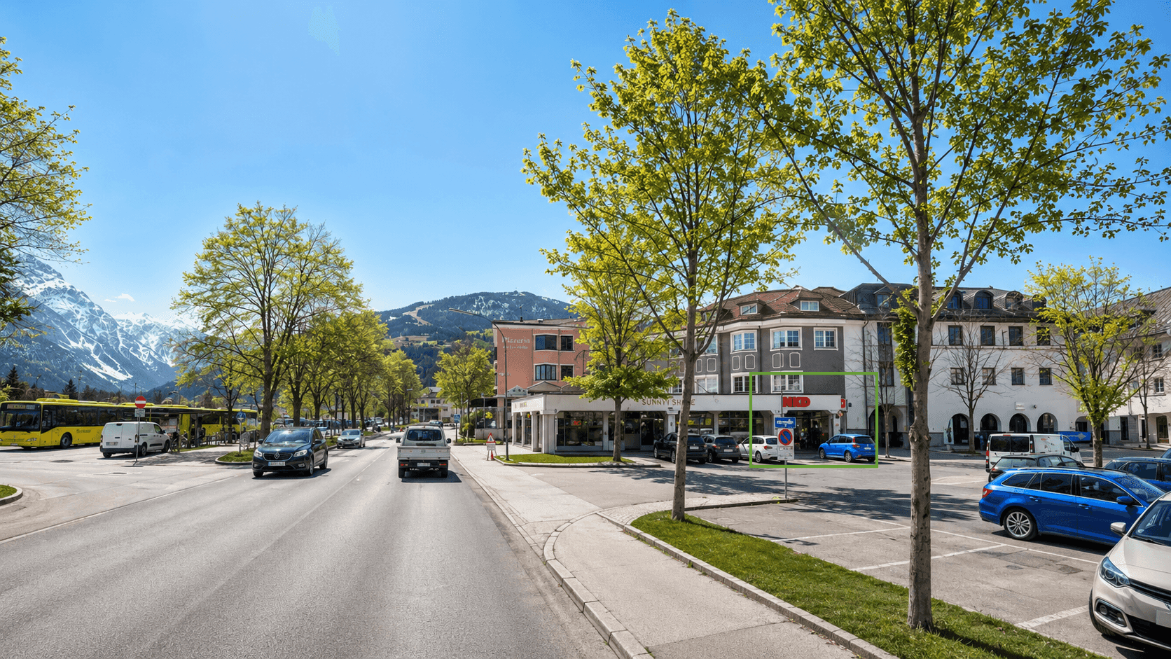 Panoramablick auf eine belebte Straße mit Geschäften, Bergen im Hintergrund und parkenden Autos.