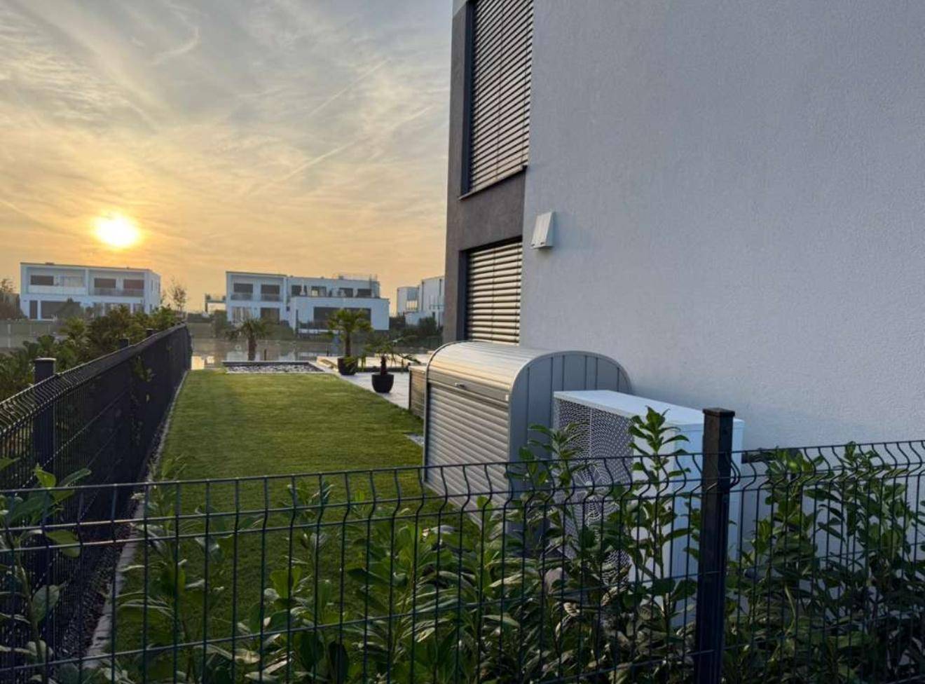 Seitenansicht des Hauses mit Garten, Zaun und Wärmepumpenanlage bei Sonnenuntergang.