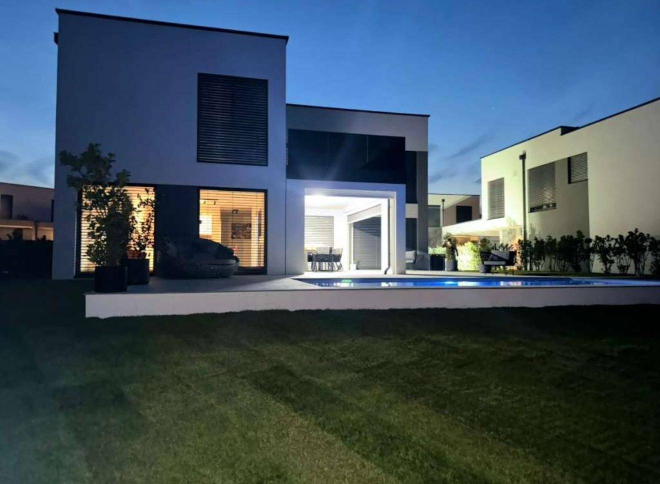 Moderne Villa mit beleuchtetem Pool und Garten bei Einbruch der Dunkelheit.