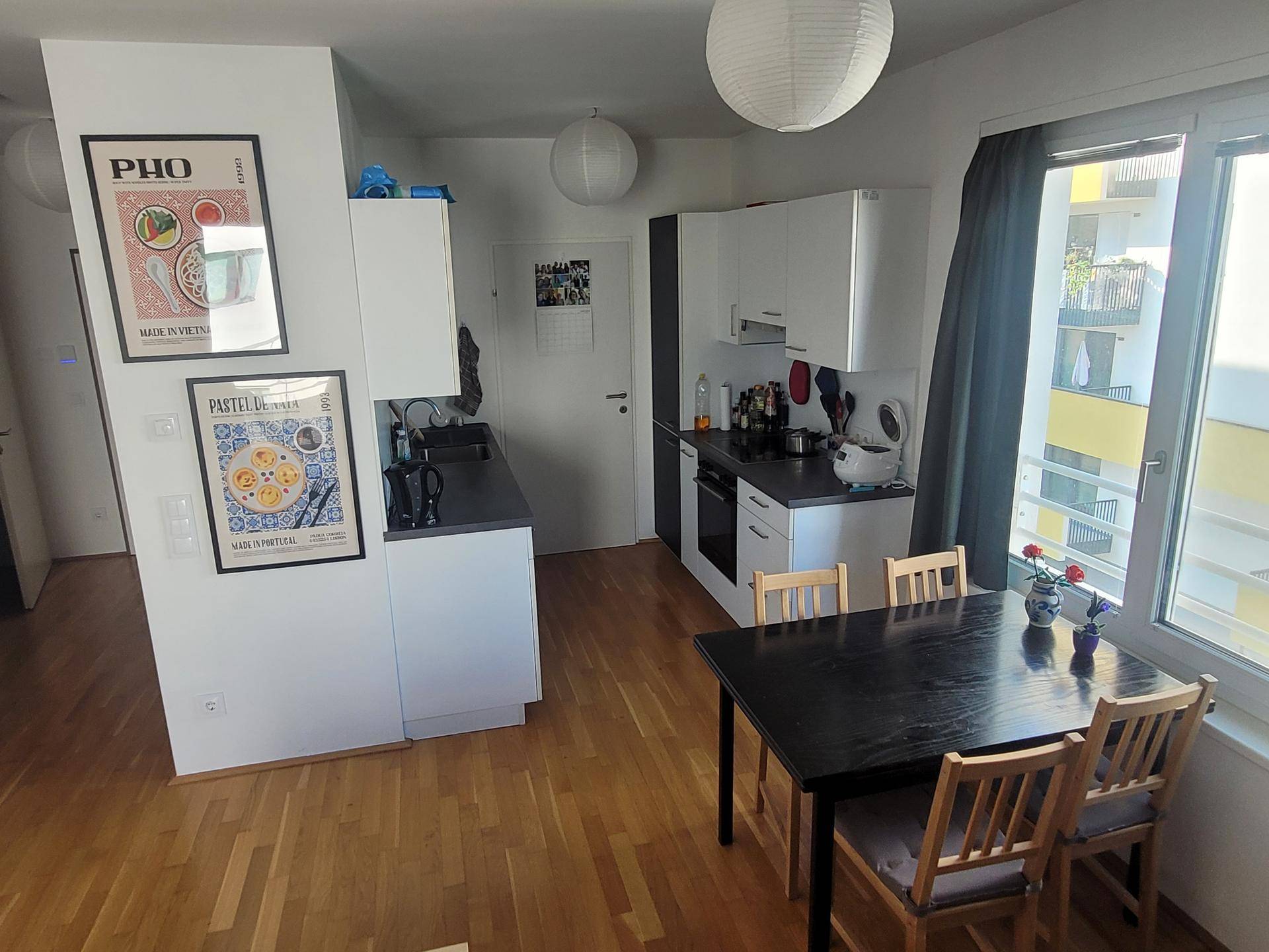 Offene Küche mit weißen Schränken, dunkler Arbeitsplatte und Essbereich am Fenster.