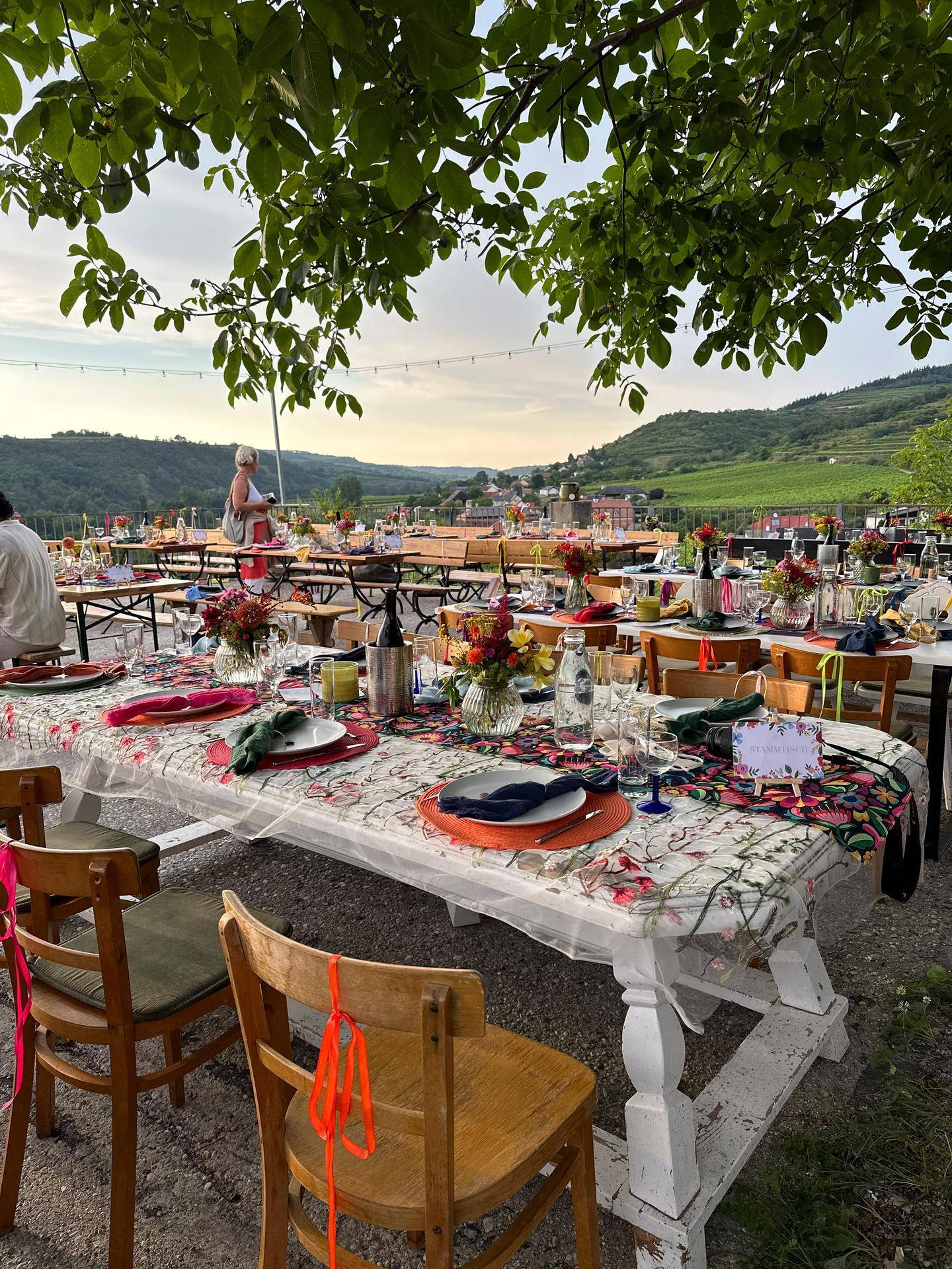 Festlich gedeckter Essbereich im Freien unter Bäumen mit Blick auf die umliegenden Hügel und ein Dorf.
