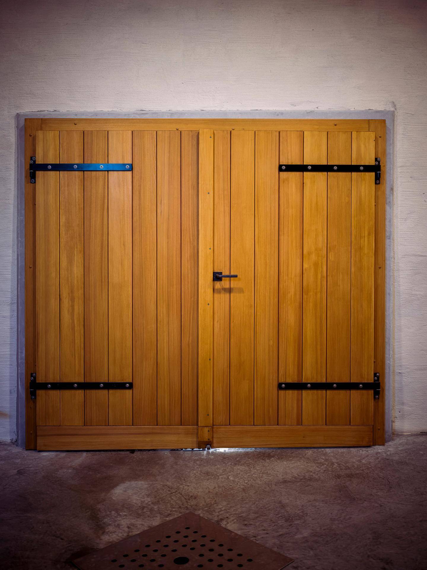 Eine große, doppelte Holztür mit schwarzen Metallscharnieren und einem Griff, eingebettet in eine helle Wand.