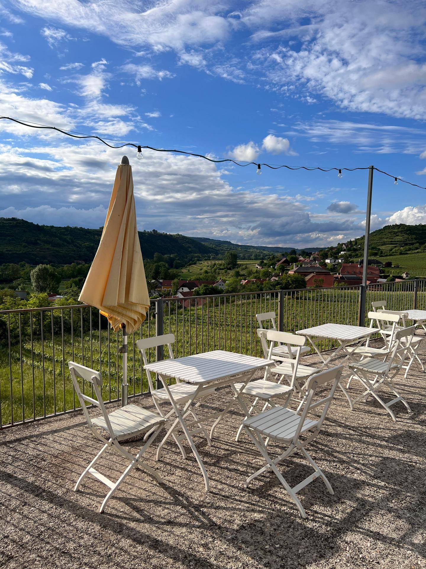 Außenterrasse mit weißen Klapptischen und Stühlen, einem Sonnenschirm und Panoramablick auf ein Tal mit Weinbergen.