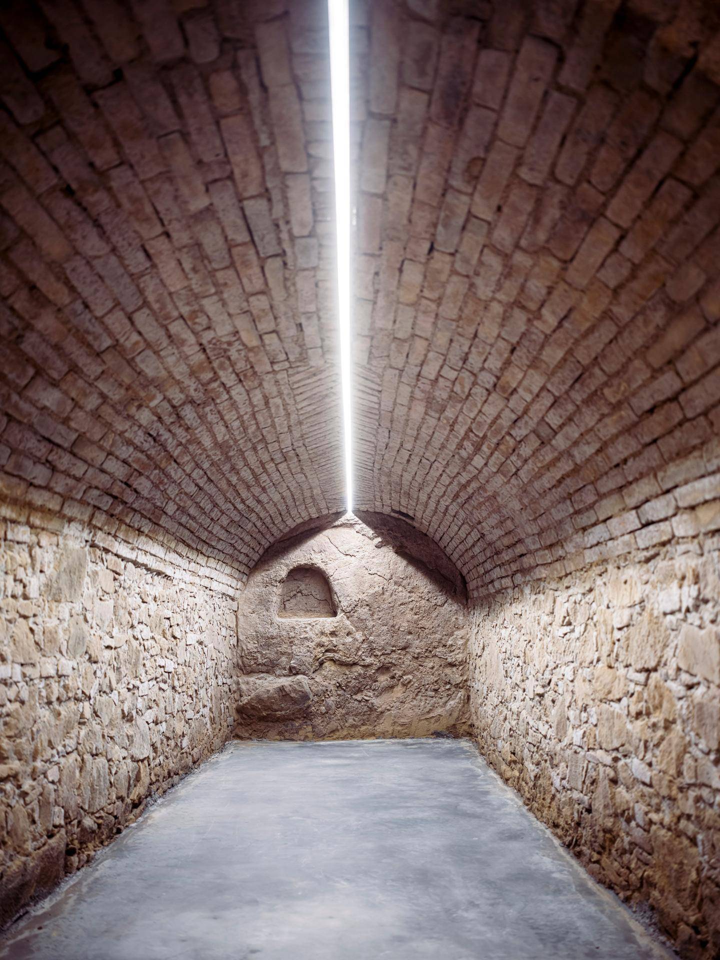 Ein langer, schmaler, gewölbter Steintunnel oder Keller mit einem einzelnen Lichtstreifen an der Decke.