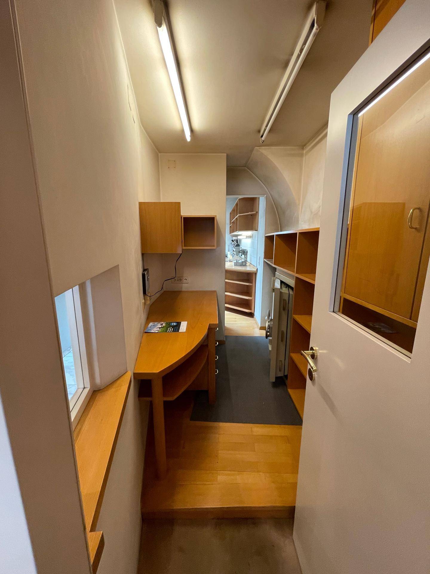 Kleiner Bürobereich mit Holzschreibtisch, Regalen und einem Fenster, ideal für administrative Aufgaben.