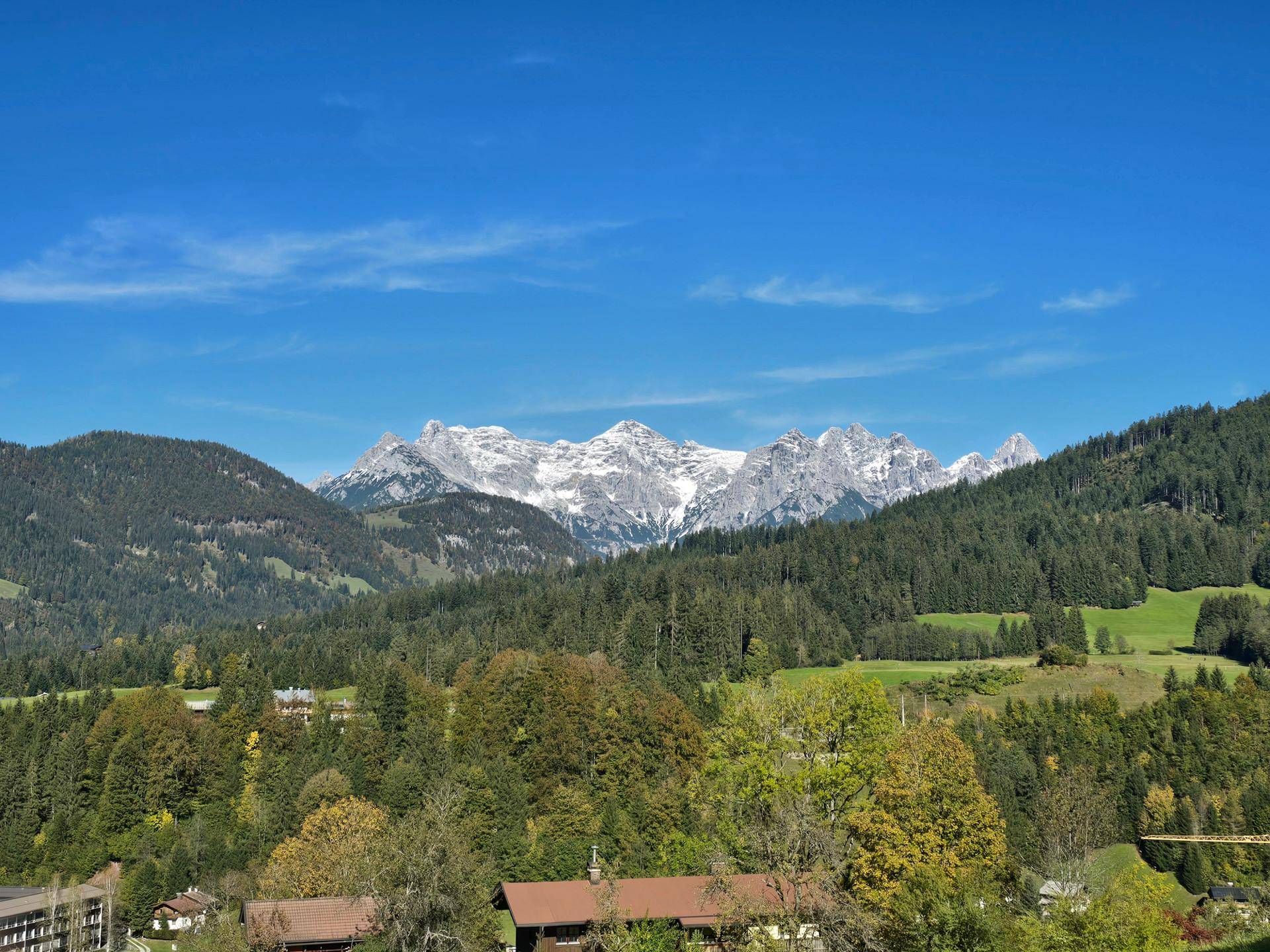 Majestätische schneebedeckte Berge ragen über dichten grünen Wäldern und sanften Hügeln unter blauem Himmel auf.