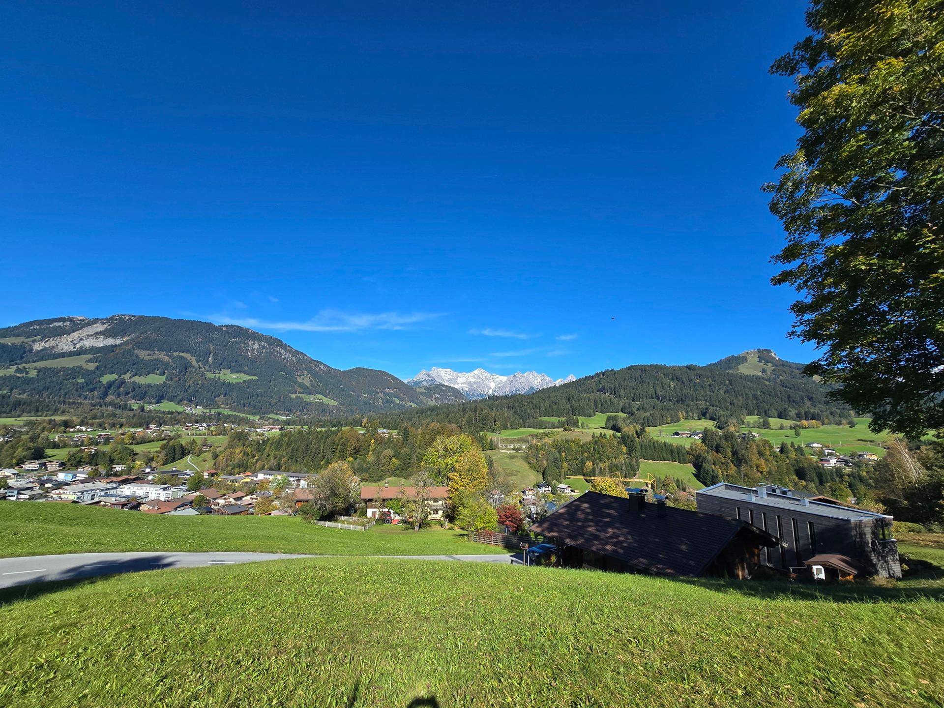 Panoramablick über grüne Hügel und ein malerisches Dorf, umgeben von bewaldeten Bergen und einem klaren blauen Himmel.
