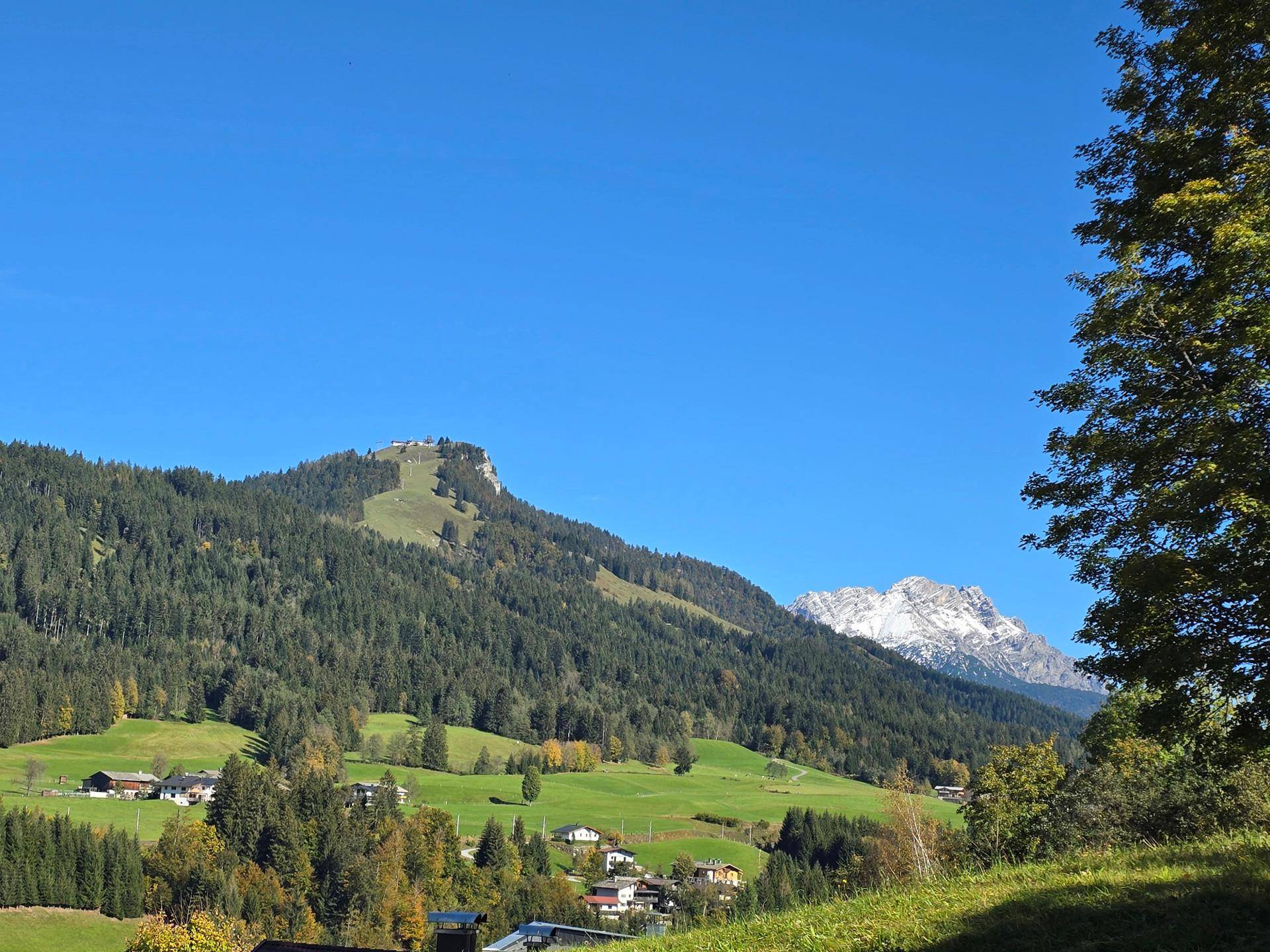 Weitläufige Alpenlandschaft mit grünen Hügeln, einem kleinen Dorf und imposanten Bergen im Hintergrund.