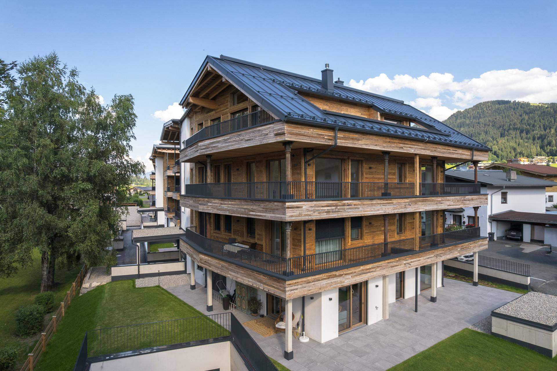 Moderne Wohnanlage mit Holzfassade, Terrassen und Gartenbereichen, umgeben von grüner Landschaft.