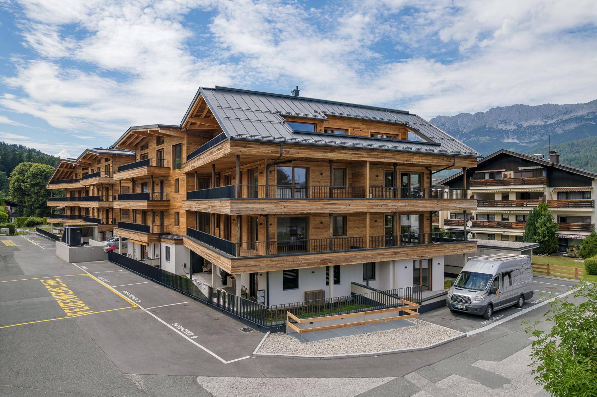 Neubau mit Holzfassade und Balkonen, umgeben von Parkplätzen und alpiner Landschaft.