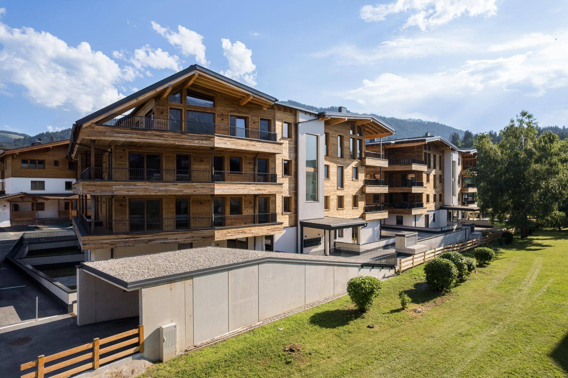 Moderne Wohngebäude mit Holzfassaden und Tiefgaragenzufahrt, umgeben von grünen Hügeln.