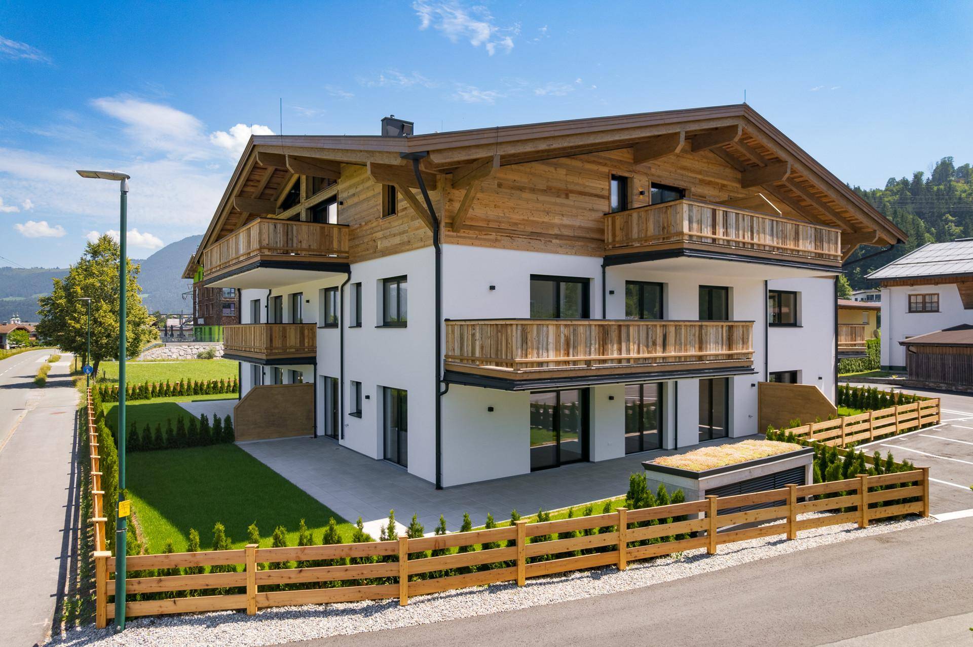 Neubau im modernen alpinen Stil mit Holzbalkonen und gepflegter Außenanlage an einer ruhigen Straße.