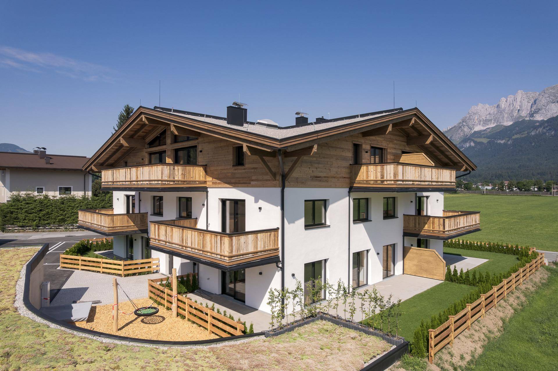 Modernes Mehrfamilienhaus im alpinen Stil mit Holzfassade, Balkonen und einem kleinen Spielplatz.