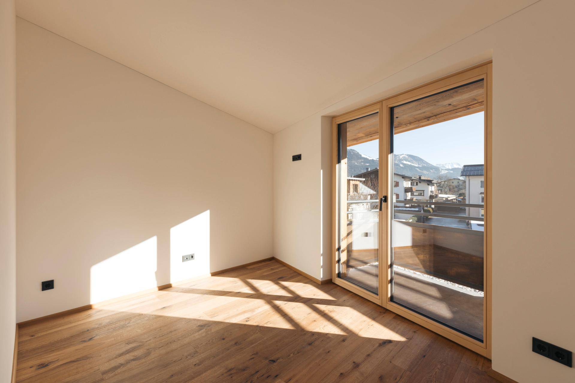Heller, leerer Raum mit Holzboden und großem Schiebefenster, das Zugang zu einem Balkon mit Bergblick bietet.