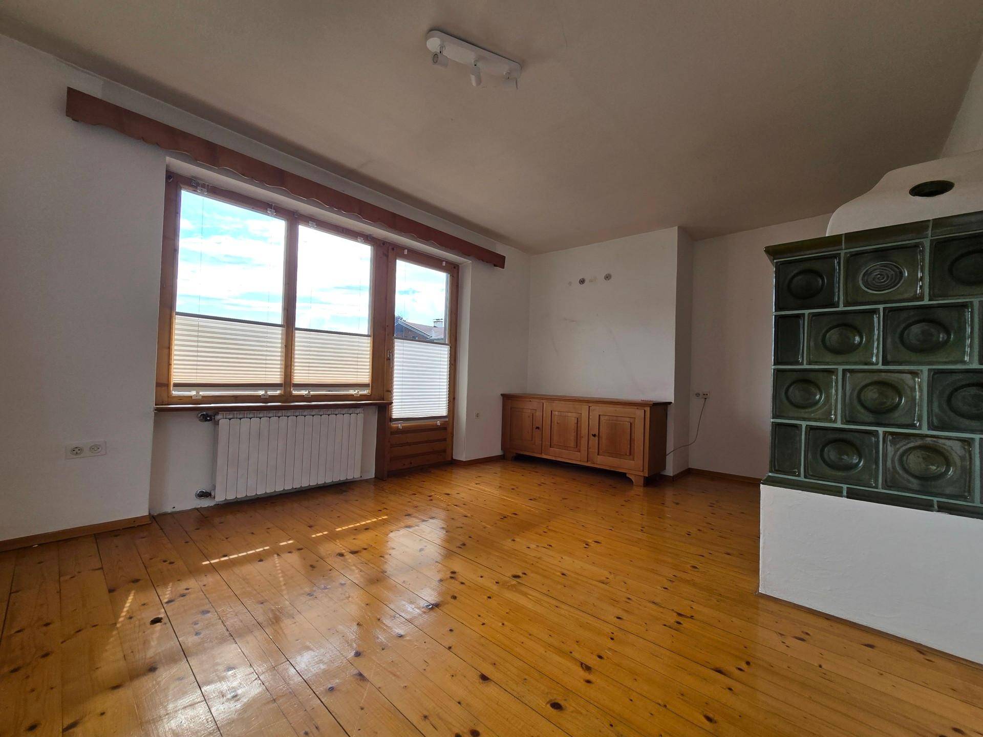 Helles Wohnzimmer mit Holzboden, großem Kachelofen und Blick ins Grüne durch die Fenster.