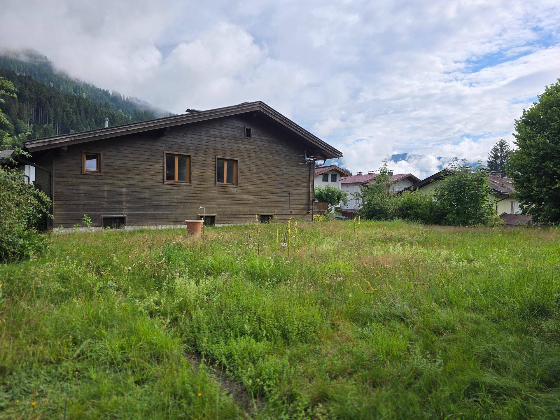Holzhaus in ländlicher Umgebung mit großer Wiese und bewaldeten Bergen im Hintergrund unter Wolken.