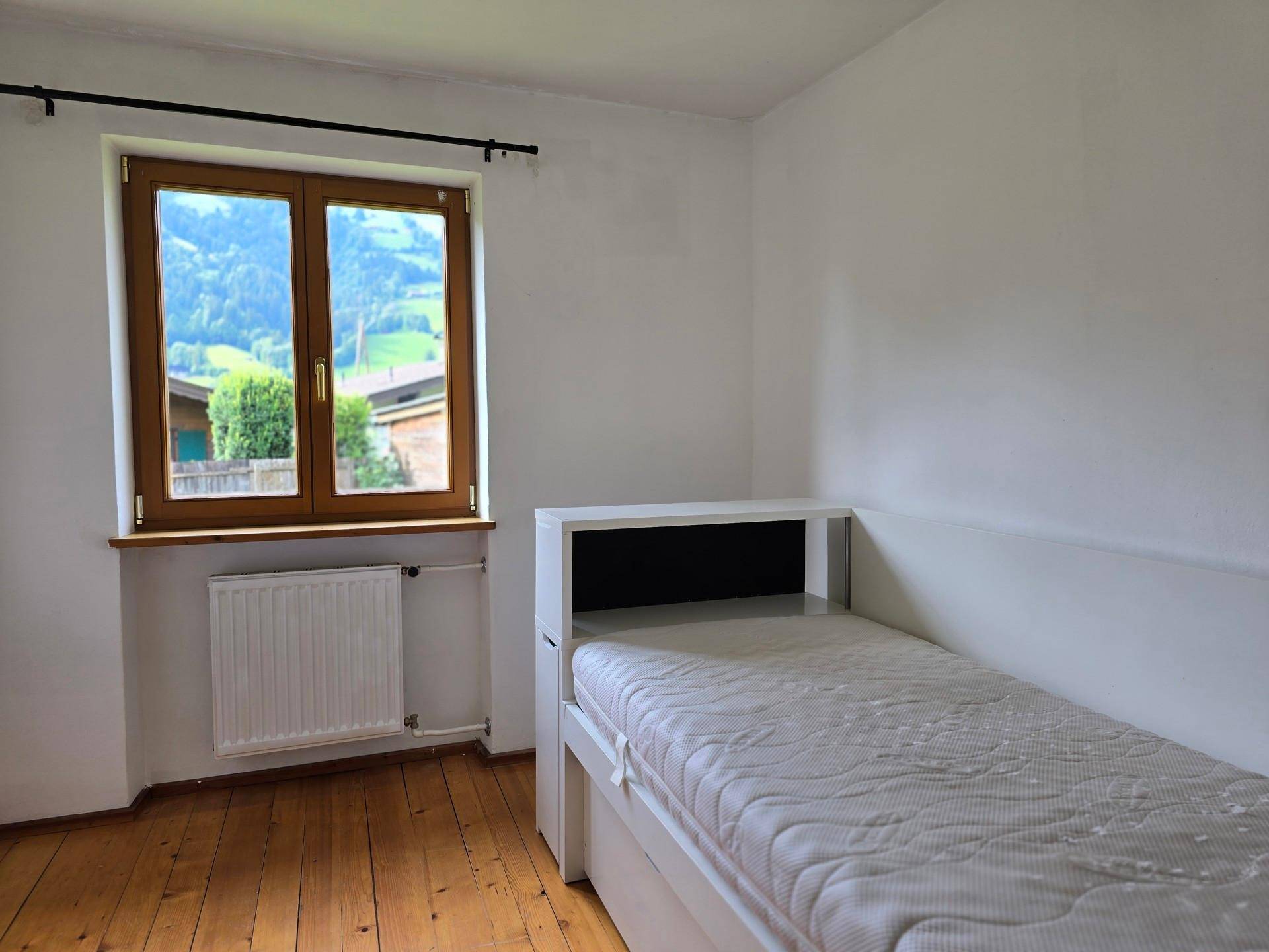 Helles Schlafzimmer mit Einzelbett, Holzboden und Fenster mit Blick auf die grünen Hügel.