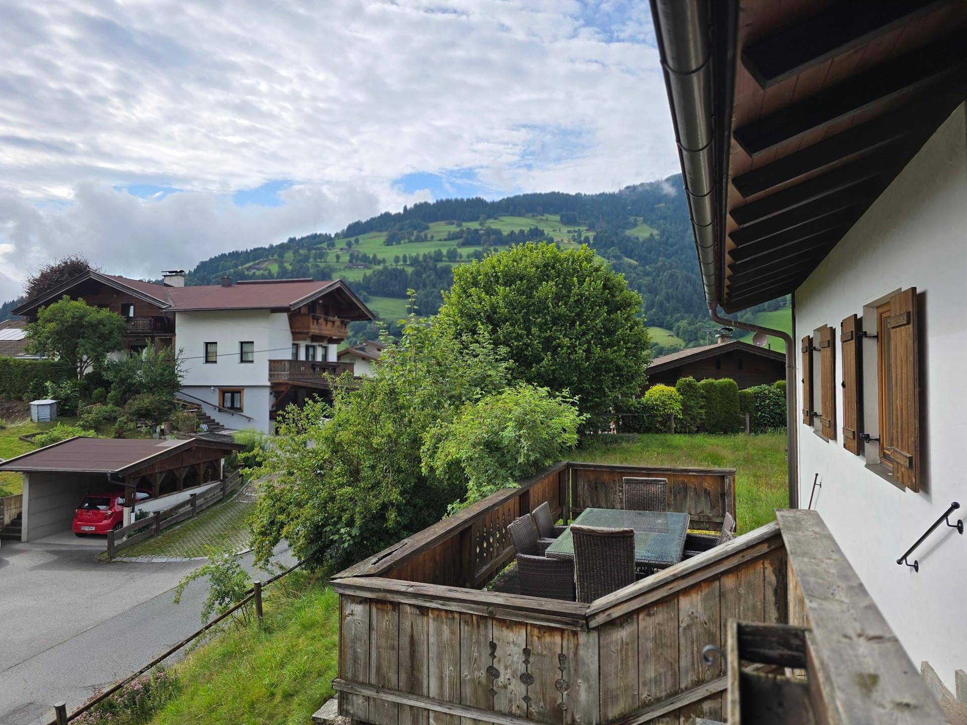 Gemütliche Holzterrasse mit Esstisch und Stühlen, umgeben von grüner Landschaft und Bergen.