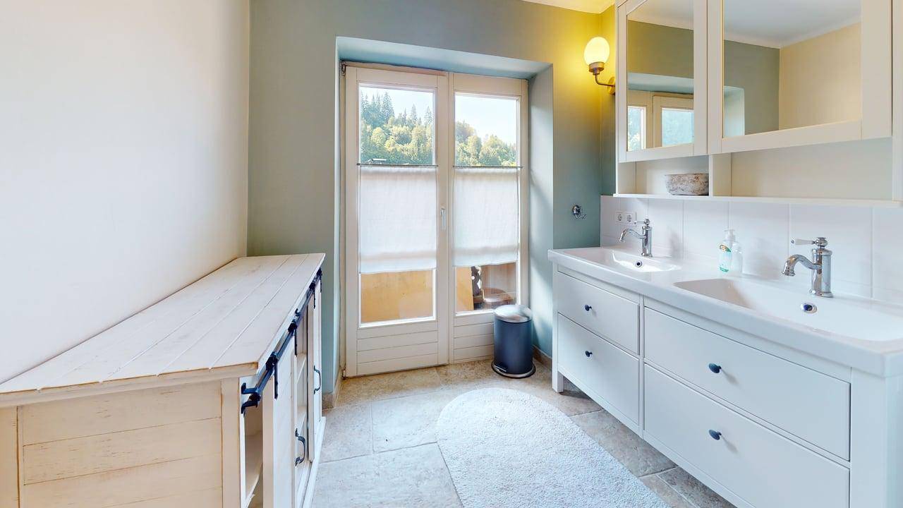 Helles Badezimmer mit Doppelwaschbecken, weißen Schränken und Fenster mit Blick ins Grüne.