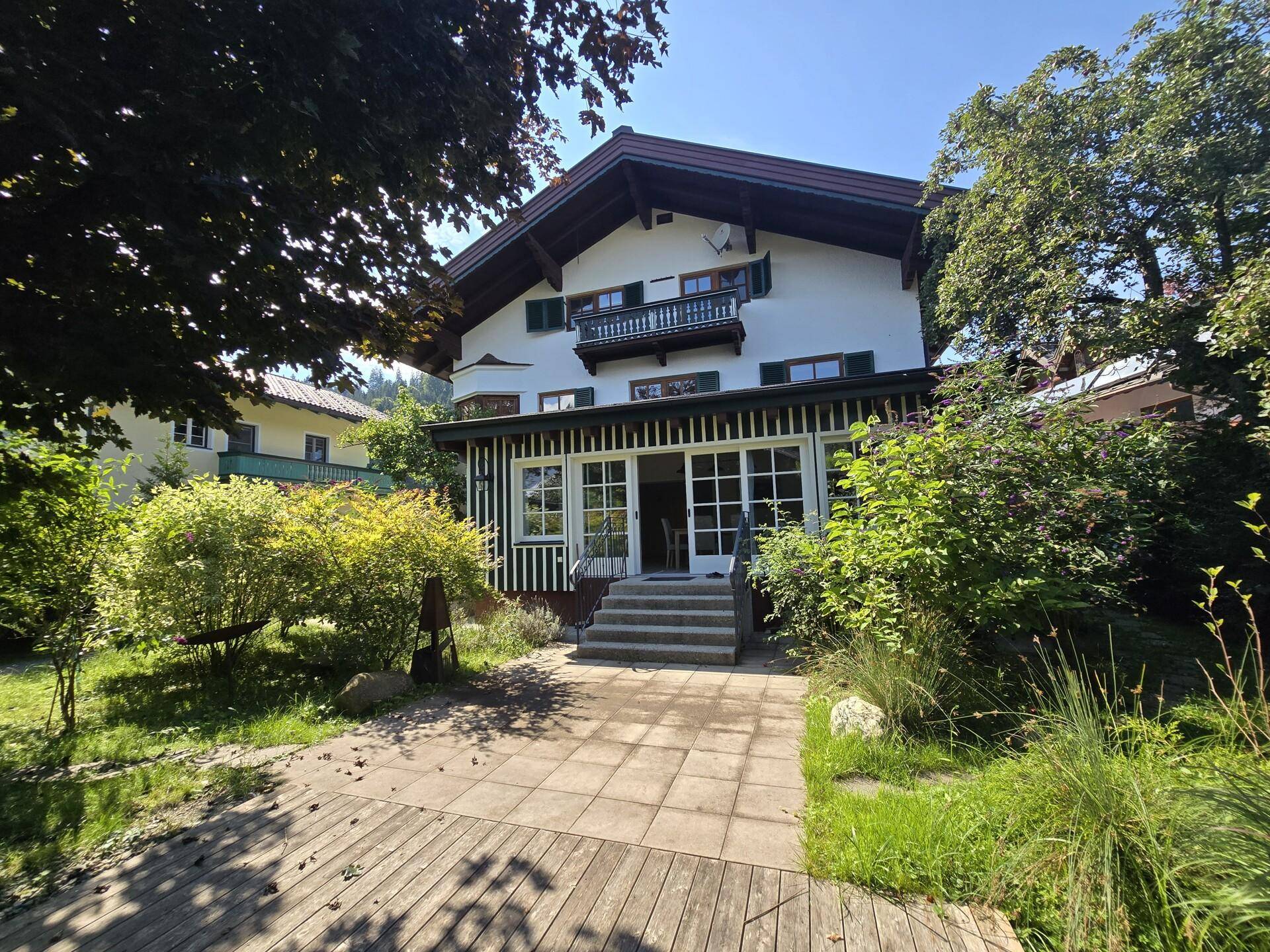 Sonniger Außenbereich mit gepflasterter Terrasse, umgeben von grünen Sträuchern und Bäumen.