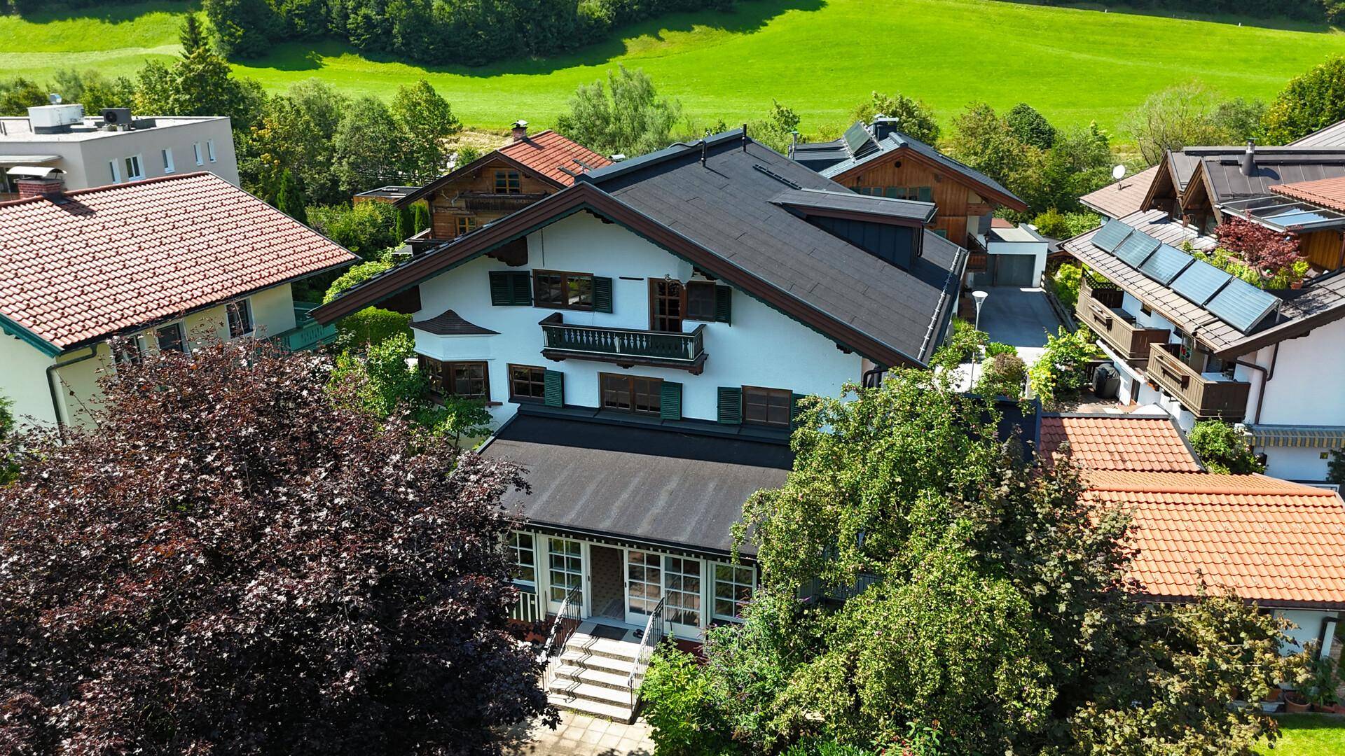 Alpenländisches Haus mit dunklem Dach und grünen Fensterläden, eingebettet in eine grüne Umgebung.
