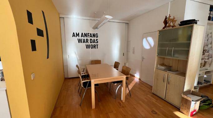 Besprechungsraum mit Holztisch und Stühlen, gelber Wand und einem Schrank mit Glasfront.