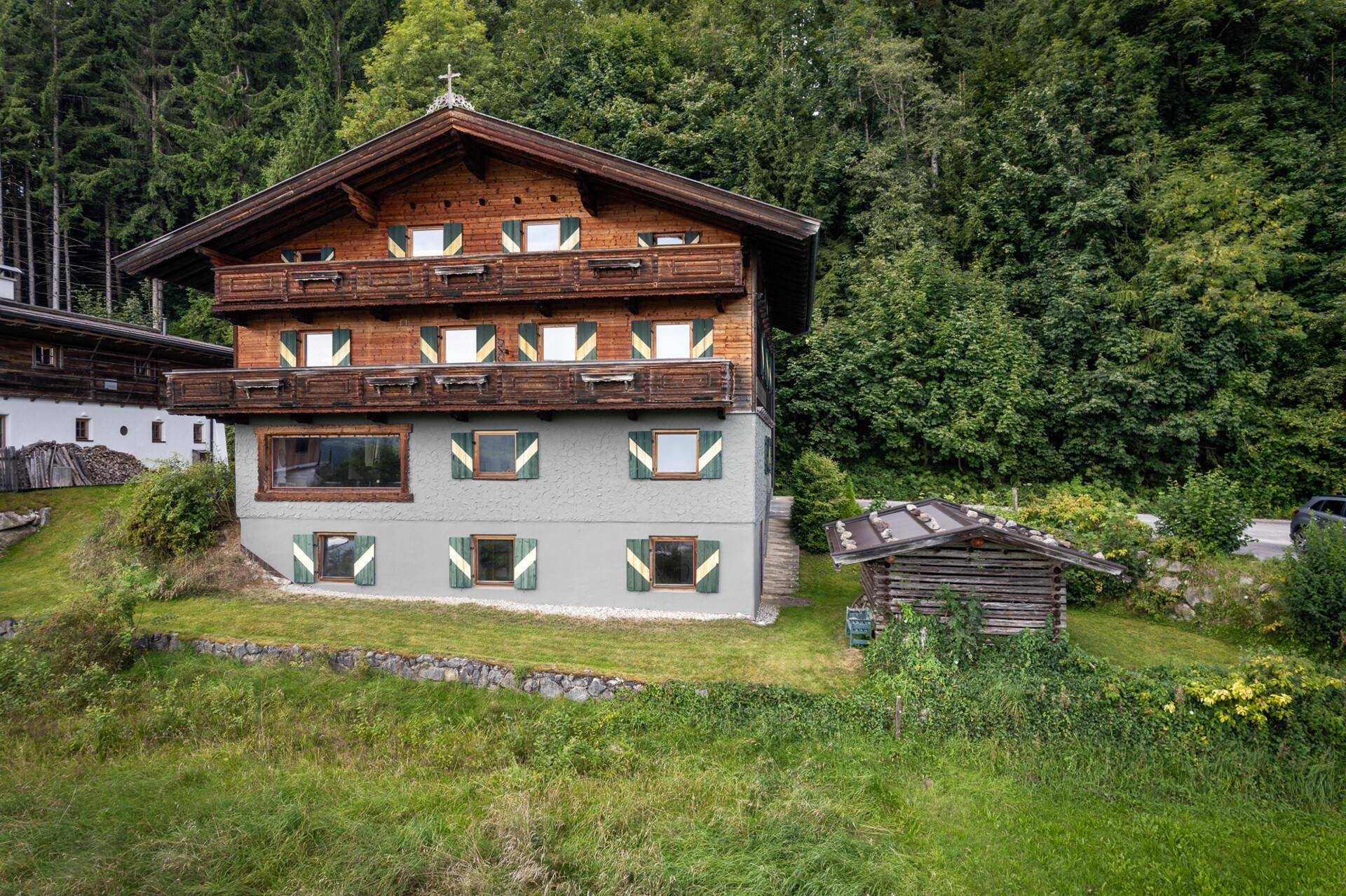 Frontale Ansicht des Alpenhauses mit Holzbalkonen und grünen Fensterläden vor bewaldetem Hintergrund.