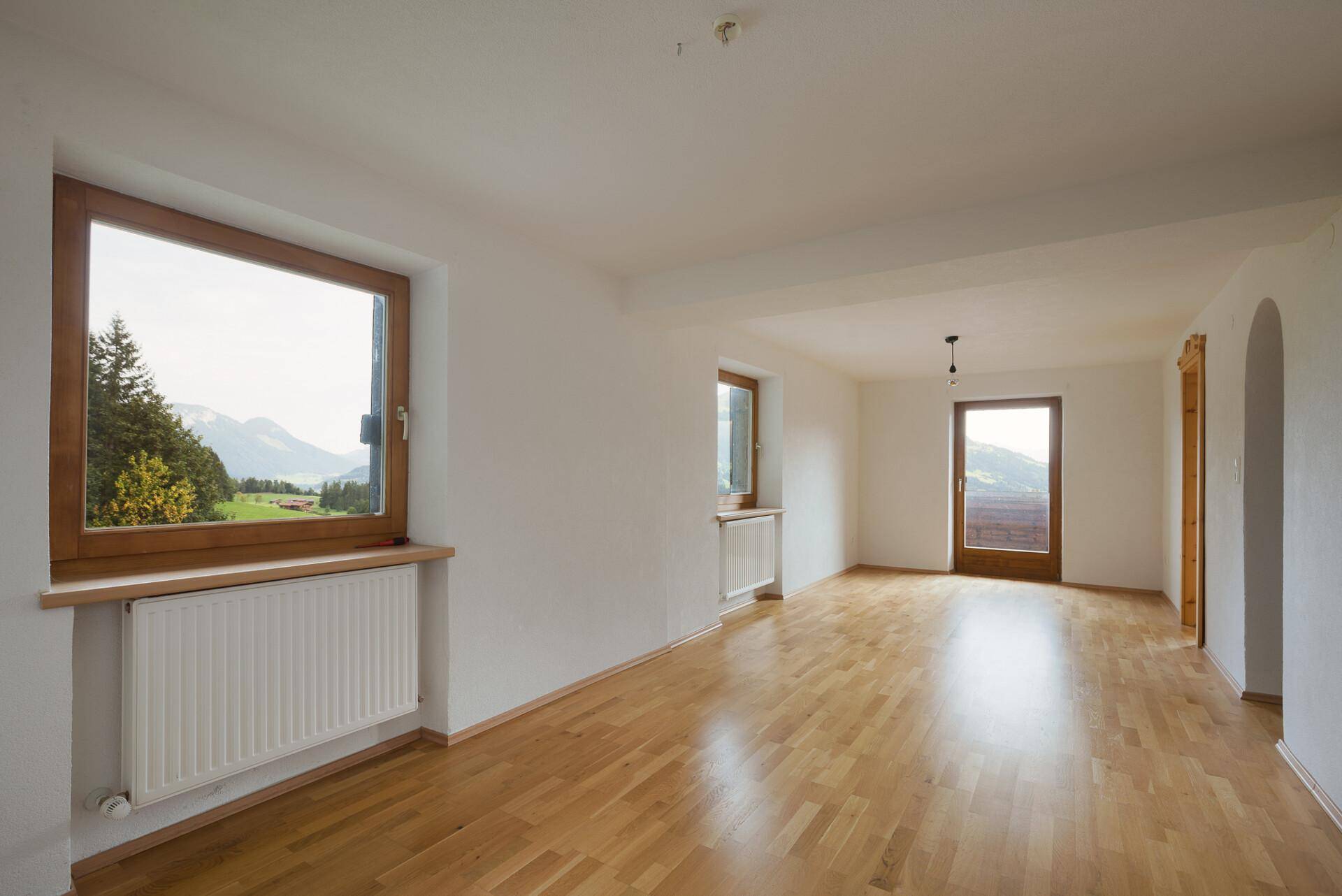 Heller, leerer Raum mit Holzfußboden, großem Fenster und weitem Blick auf die Berglandschaft.
