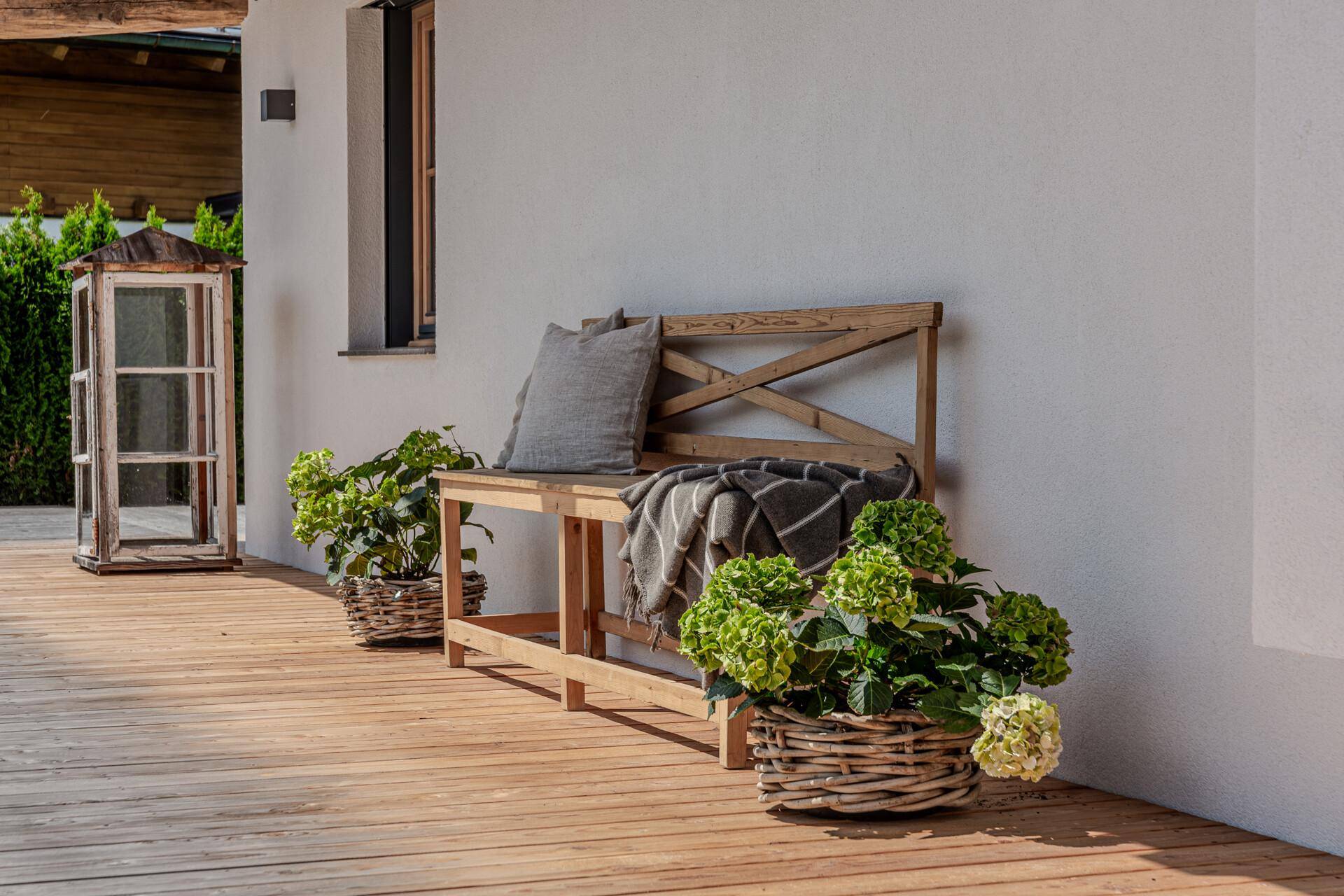 Rustikale Holzbank auf einer sonnigen Holzterrasse, dekoriert mit grünen Pflanzen in Weidenkörben.