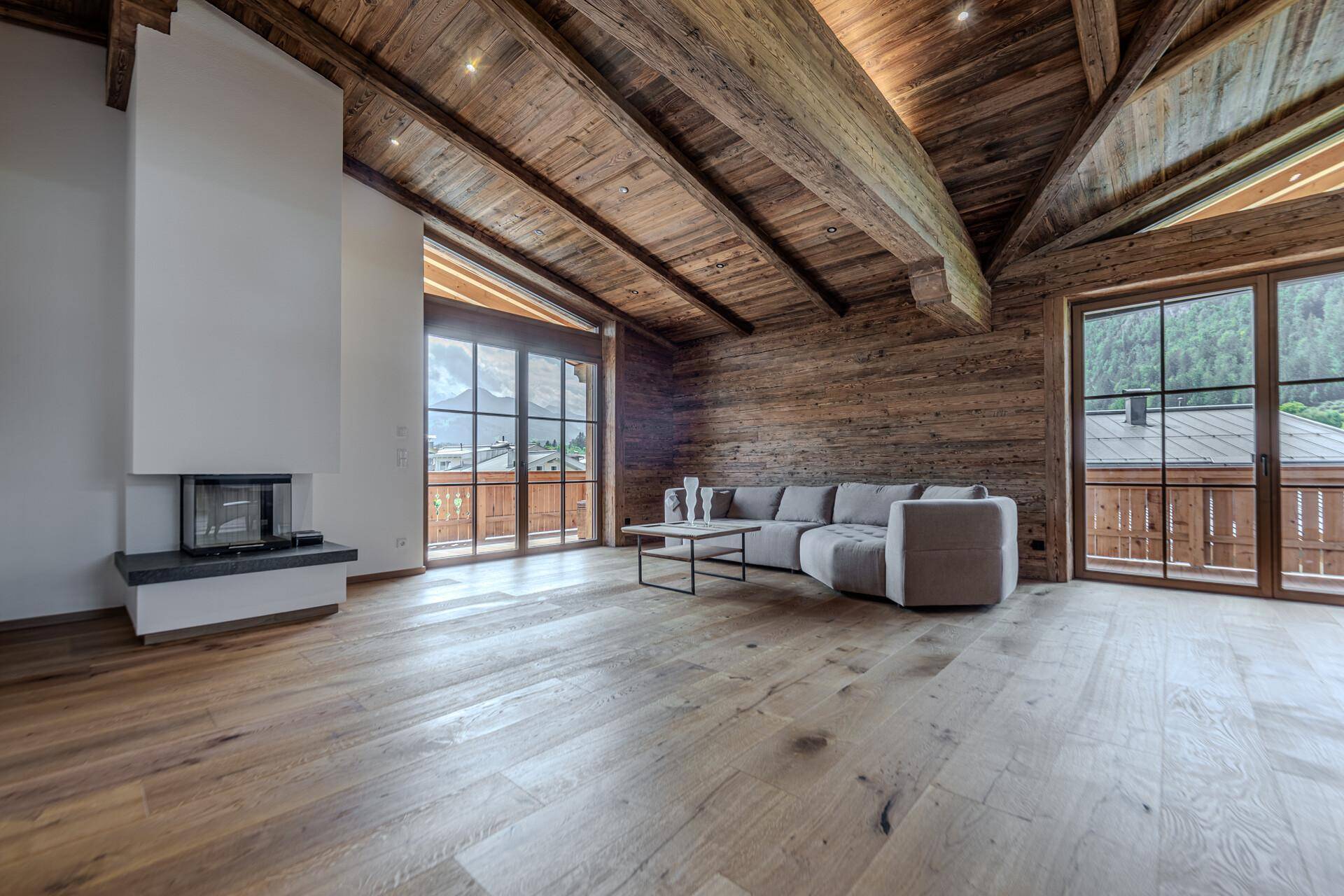 Gemütliches Wohnzimmer mit modernem Kamin, beeindruckender Holzbalkendecke und großen Fenstern mit Bergblick.
