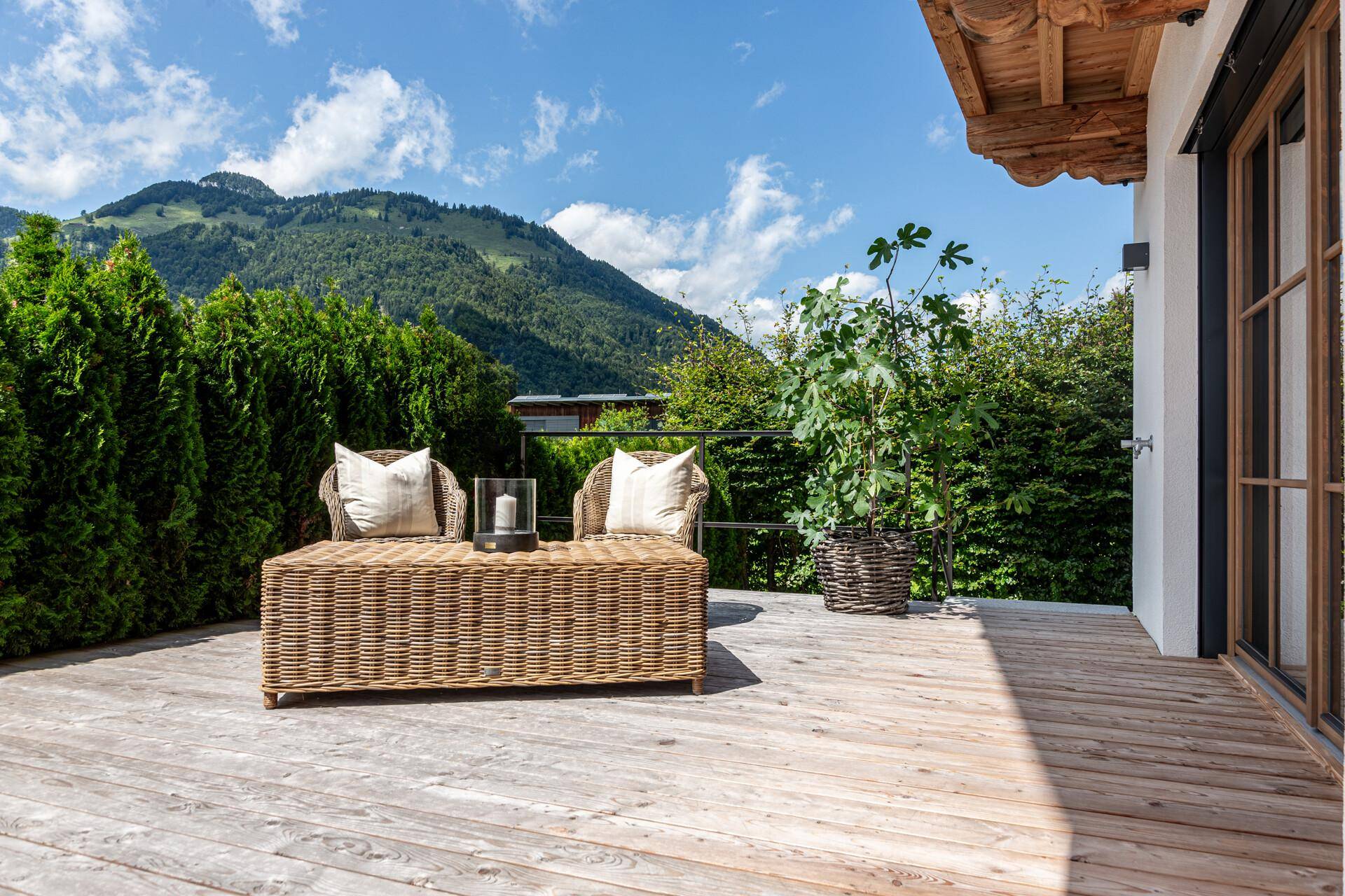 Sonnige Holzterrasse mit gemütlichen Korbmöbeln und weitem Blick auf die umliegende Berglandschaft.