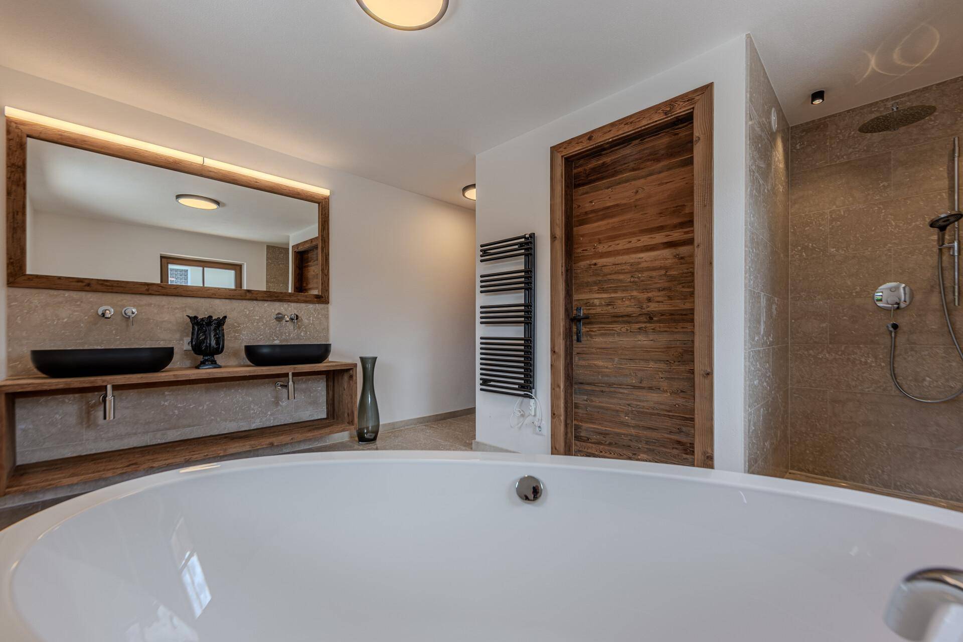 Elegantes Badezimmer mit freistehender Badewanne, Doppelwaschbecken, beheiztem Handtuchhalter und Holztür.