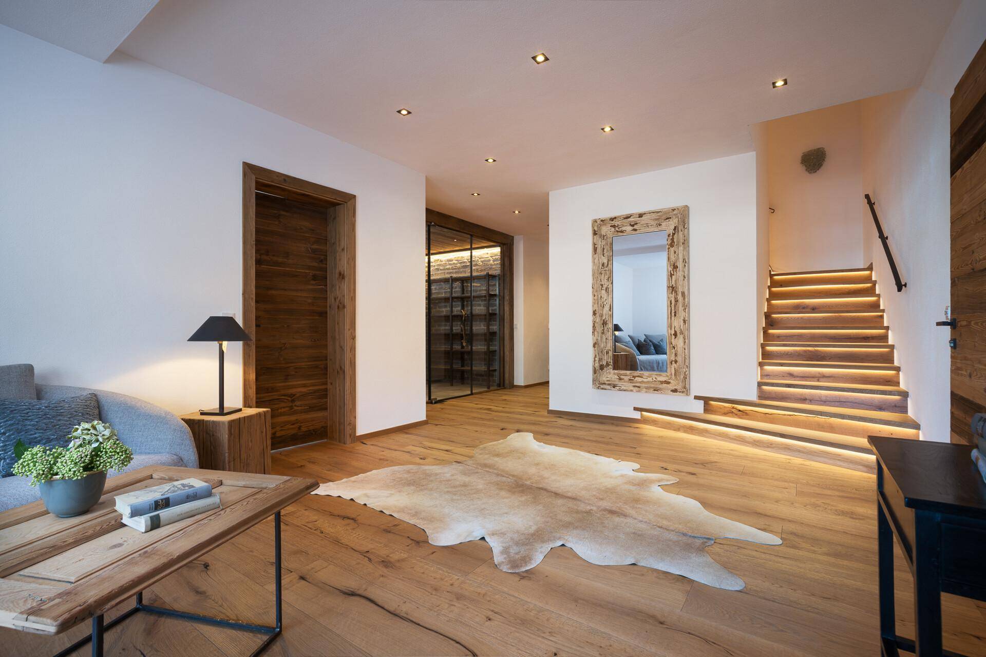 Eleganter Flur mit beleuchteter Holztreppe, Holzboden und einem stilvollen Weinkeller hinter Glas.