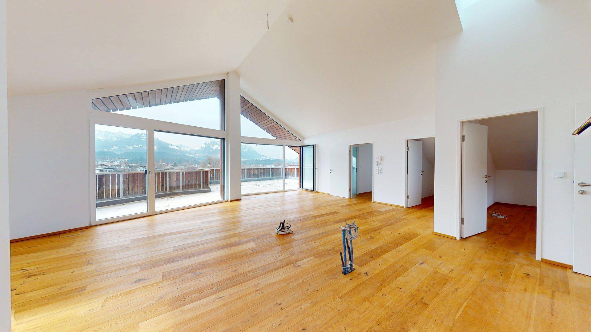 Lichtdurchfluteter Wohnraum mit Holzboden, hohen Decken und Zugang zur Terrasse mit Bergblick.