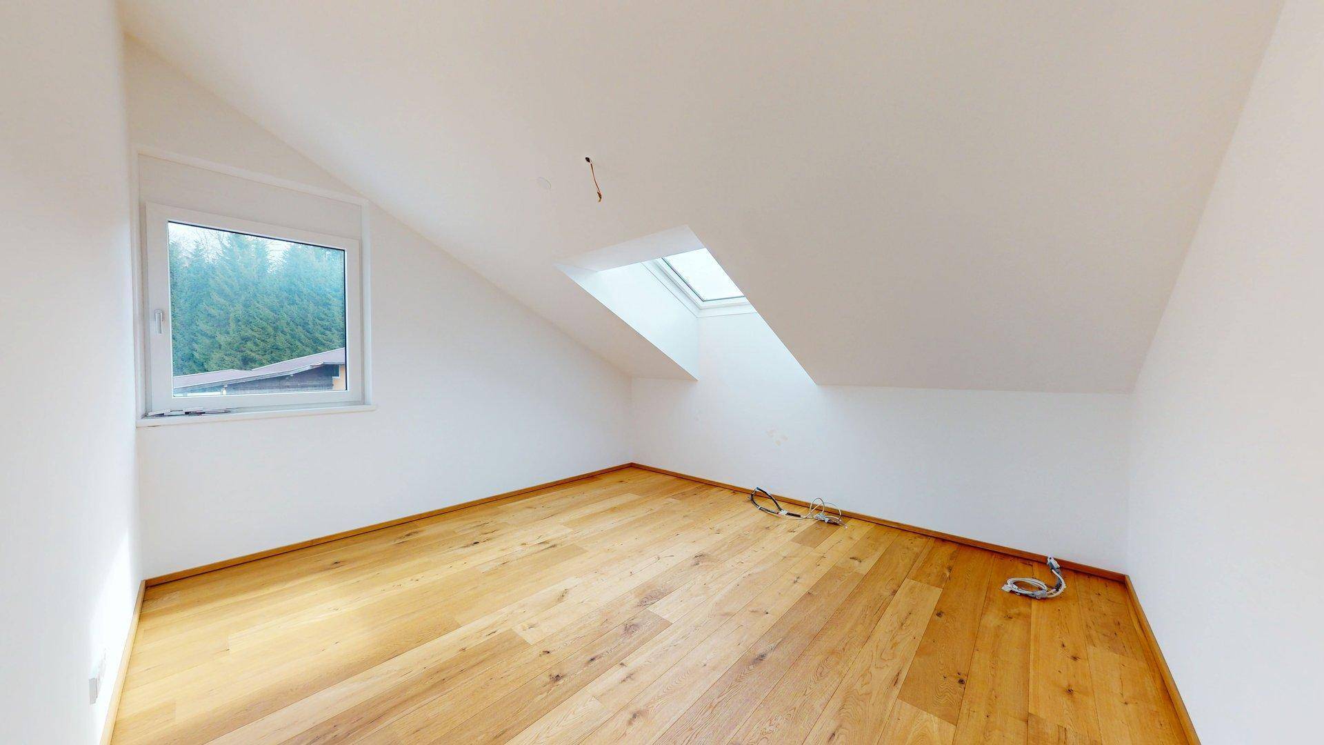 Helles Zimmer mit Holzboden, Dachschräge und Fenster mit Blick ins Grüne.
