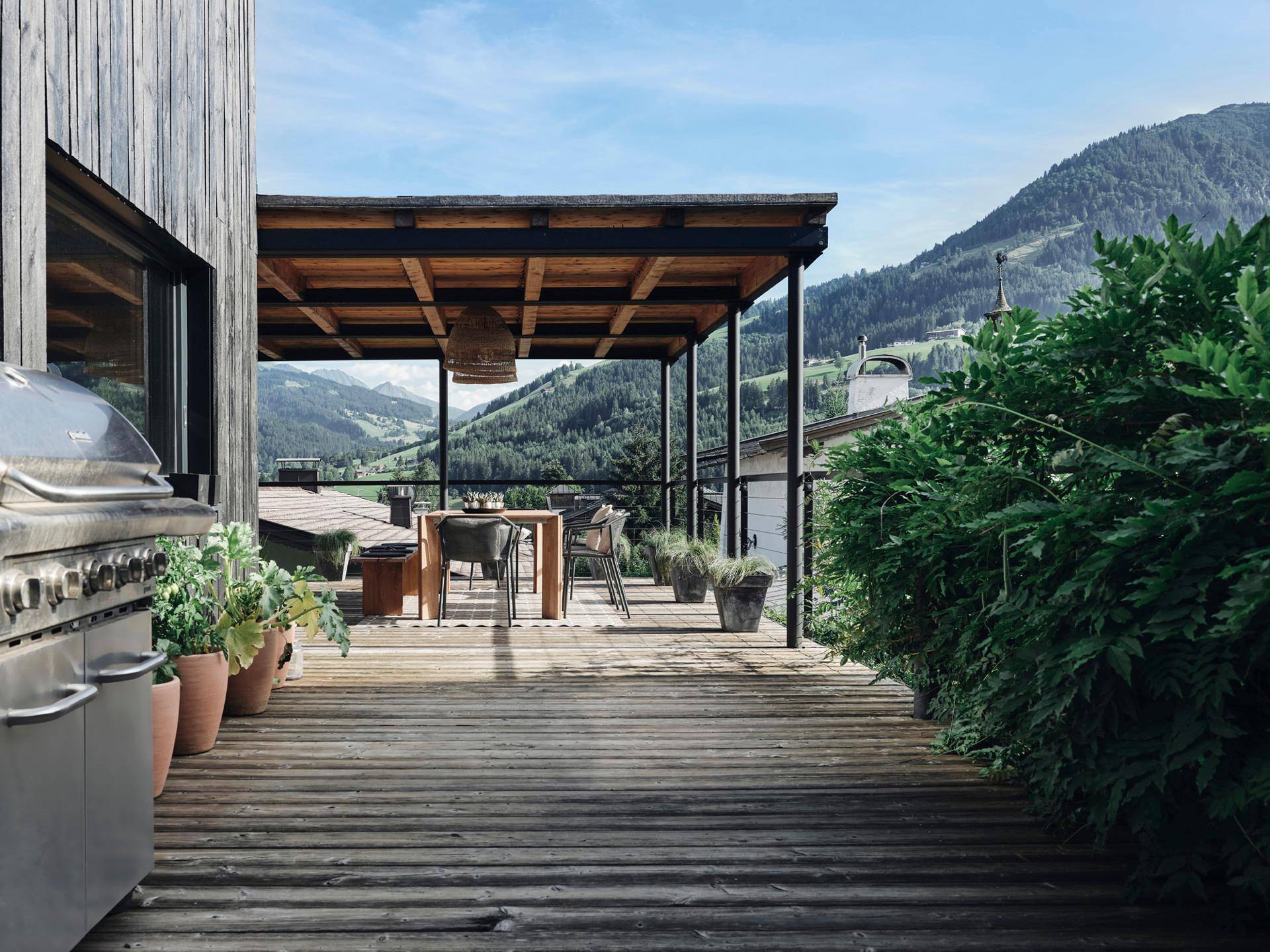 Überdachte Holzterrasse mit Grillbereich und Esstisch, umgeben von üppiger Natur und Bergblick.