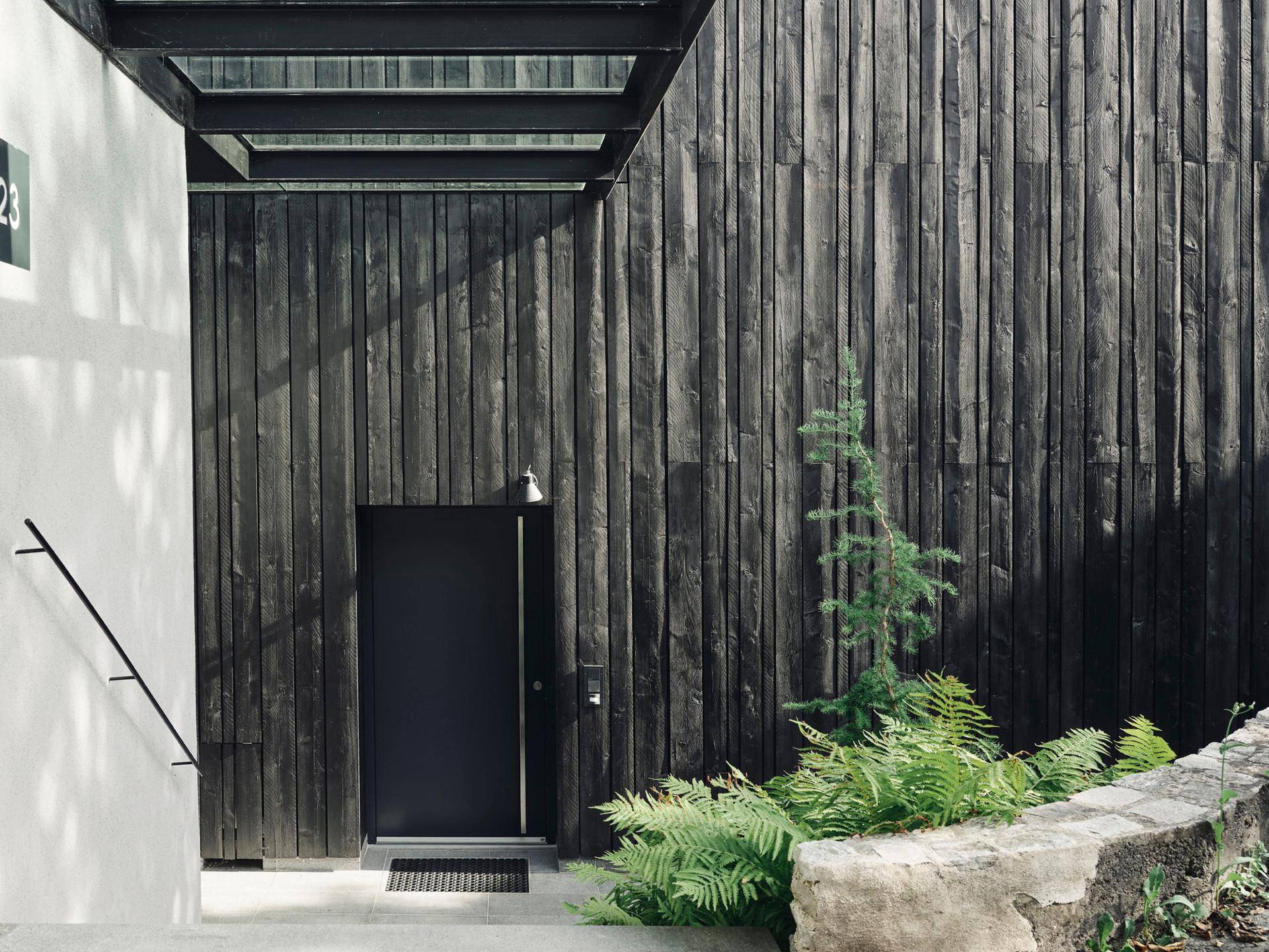 Moderner Eingangsbereich mit dunkler Holzfassade und minimalistischem Design, umgeben von Pflanzen.