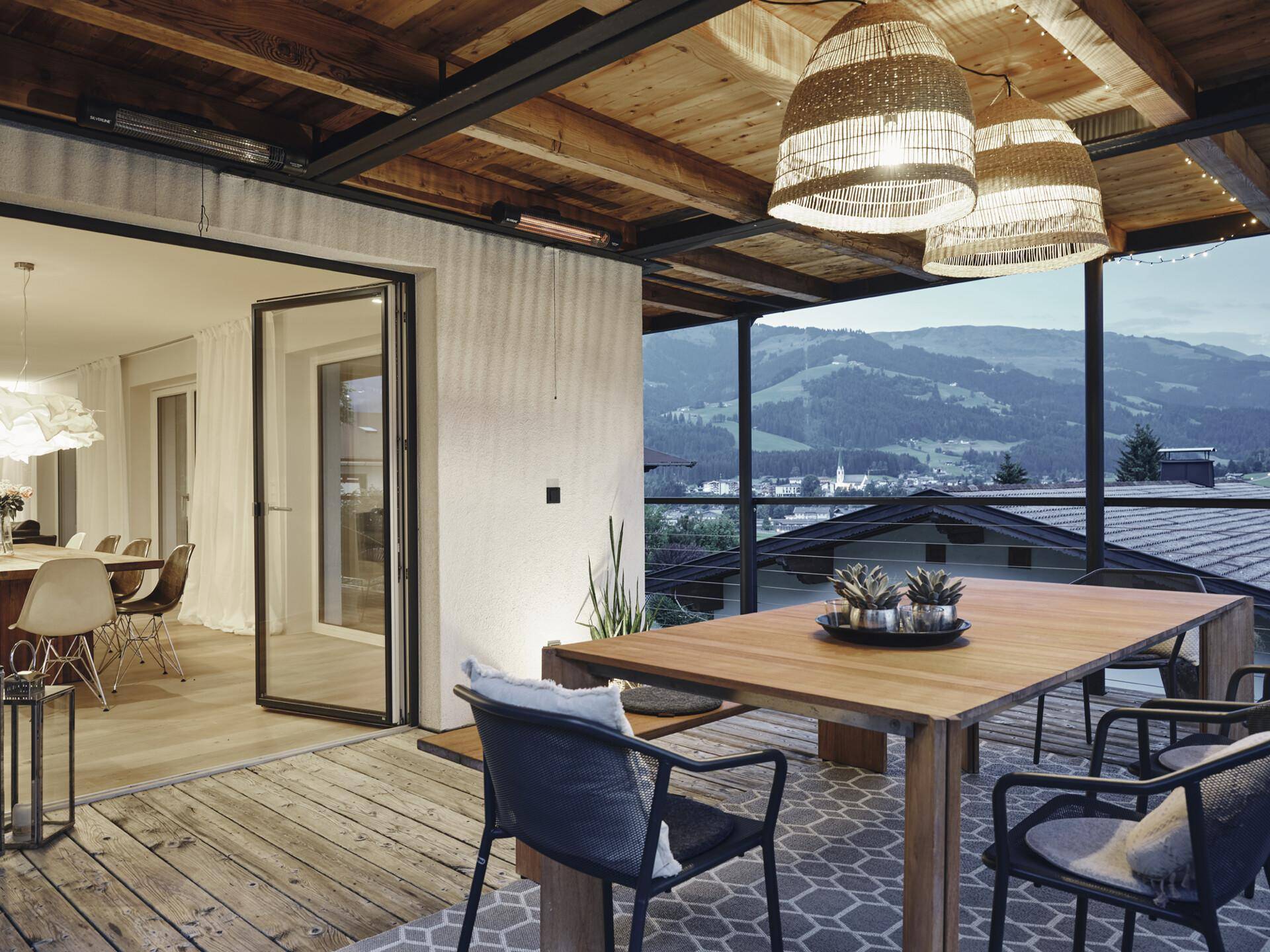 Stilvolle überdachte Terrasse mit Holzboden und Blick auf die beleuchtete Berglandschaft am Abend.