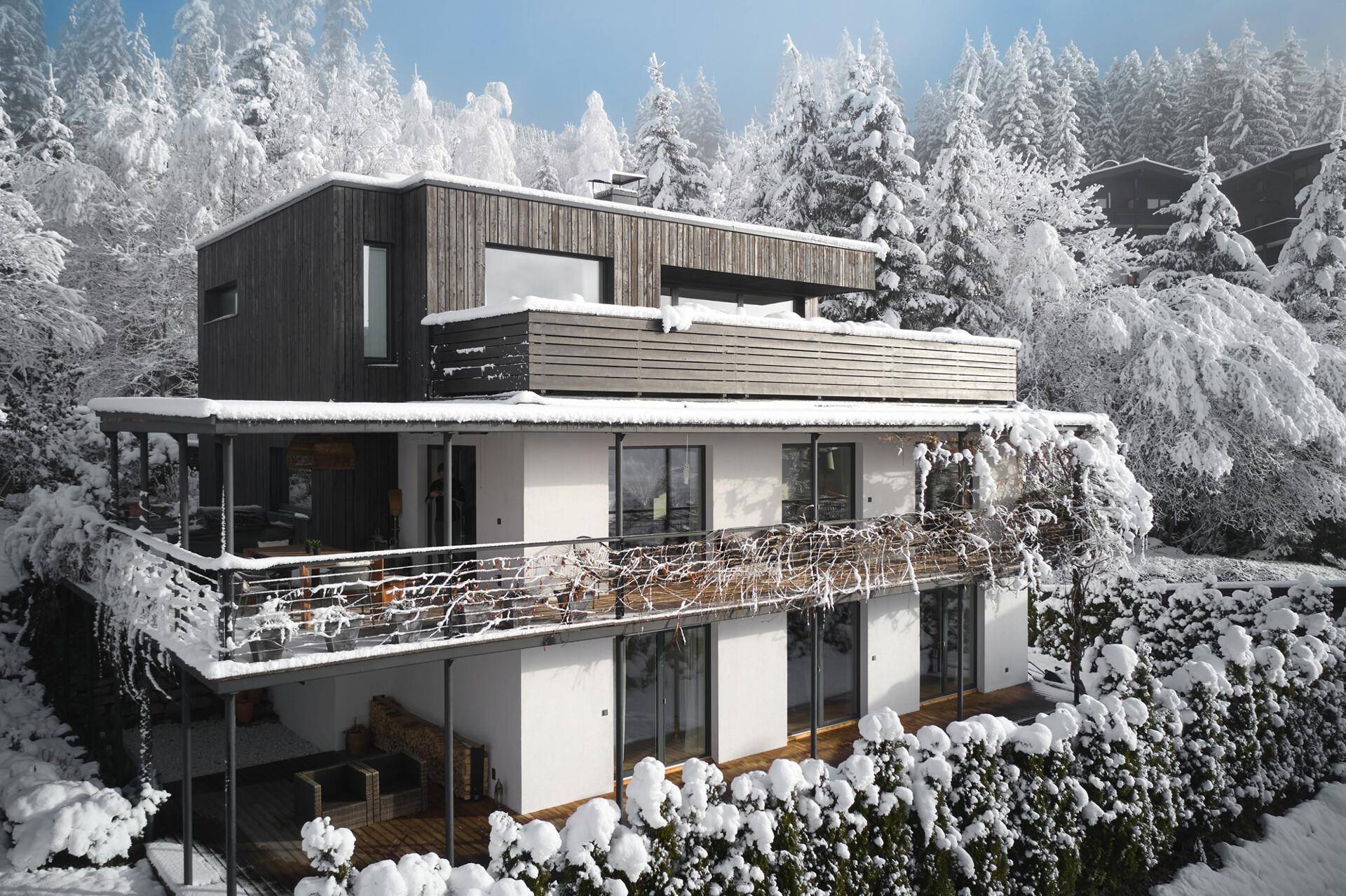 Modernes Wohnhaus im Winter, umgeben von verschneiten Bäumen und einer idyllischen Berglandschaft.