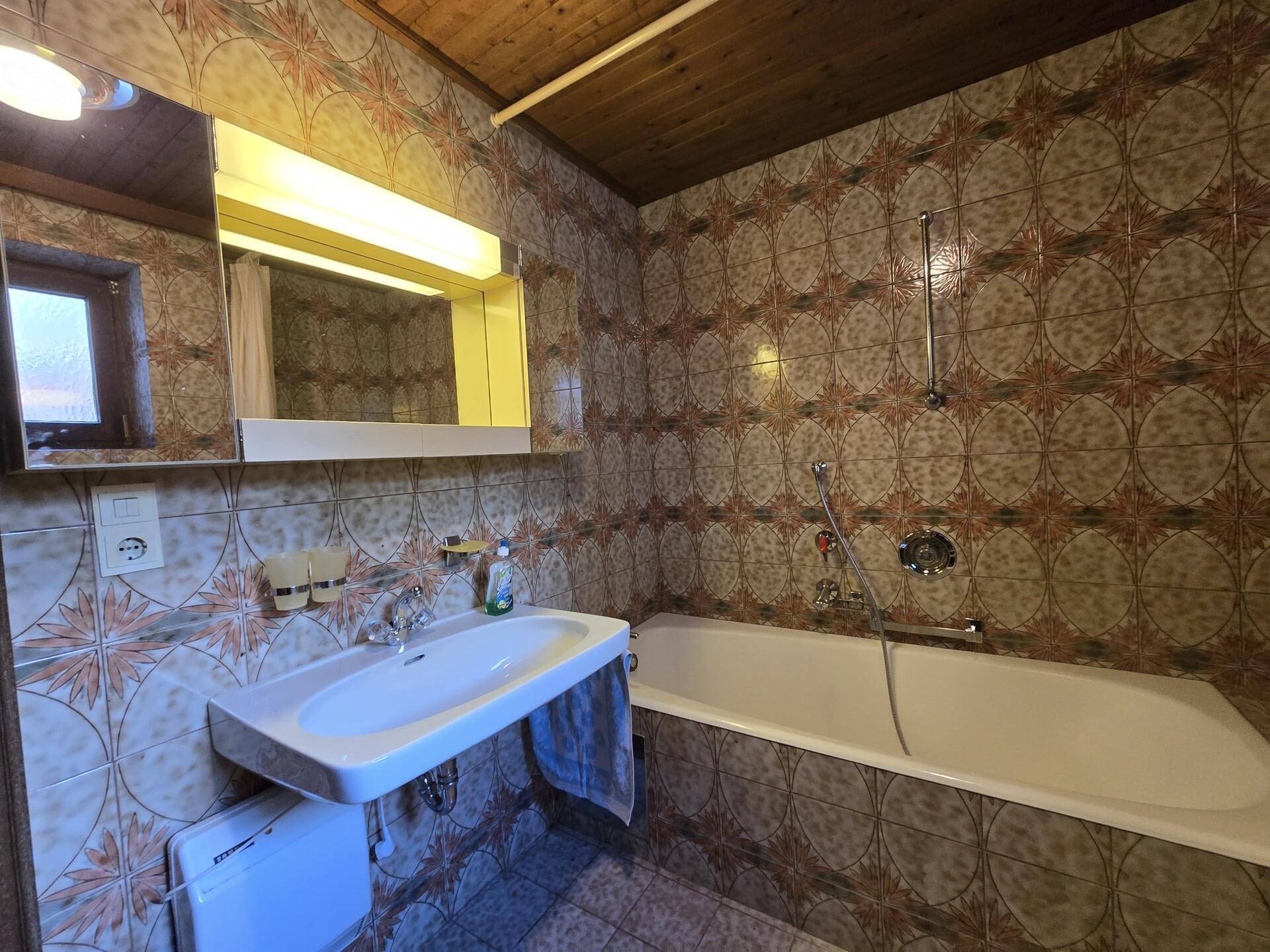 Badezimmer im 80er-Jahre-Stil mit Badewanne, Waschbecken und gemusterten Fliesen.
