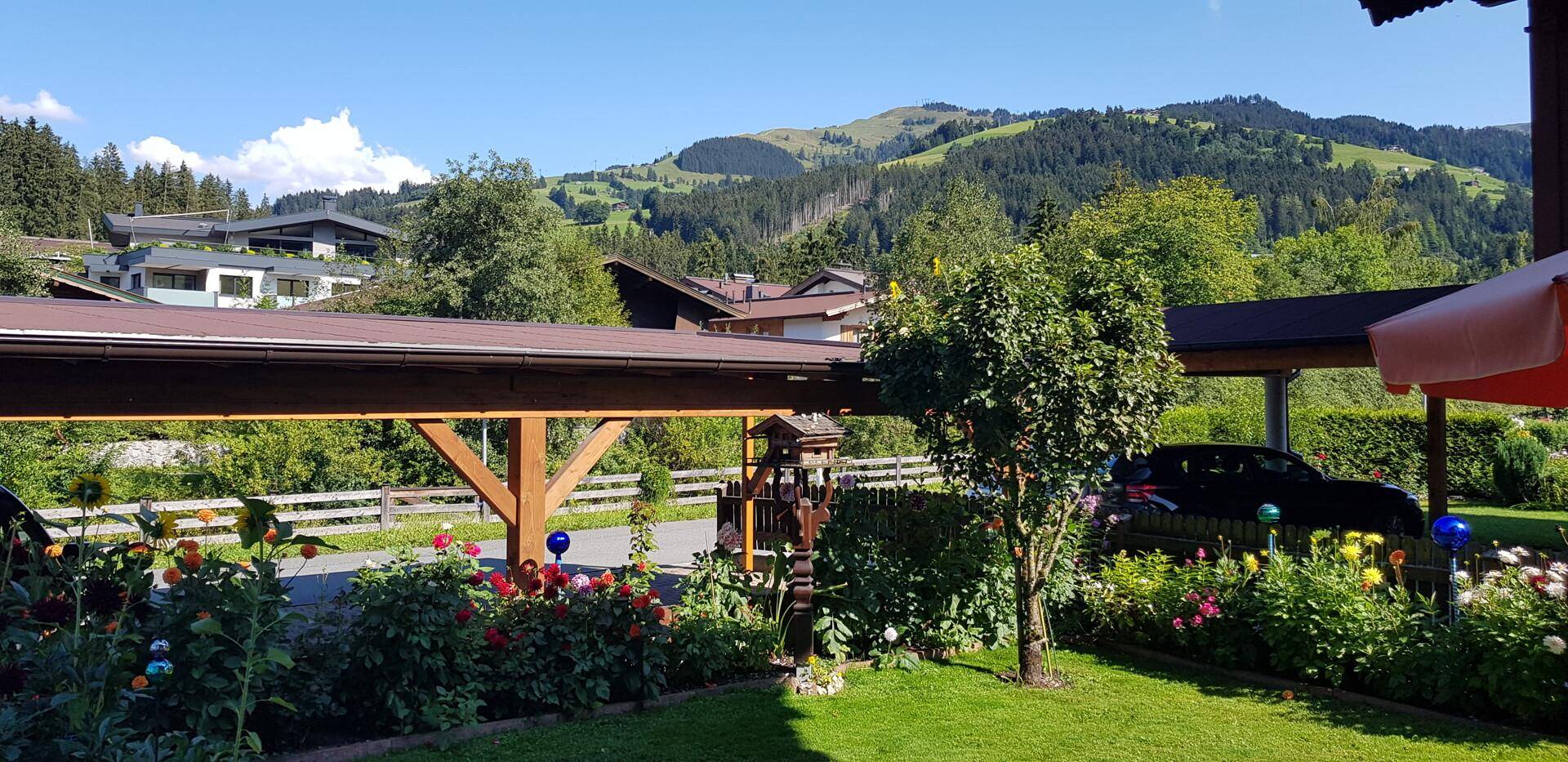 Gepflegter Garten mit Rasenfläche, Blumenbeeten und einem Carport im Hintergrund, umgeben von Natur.