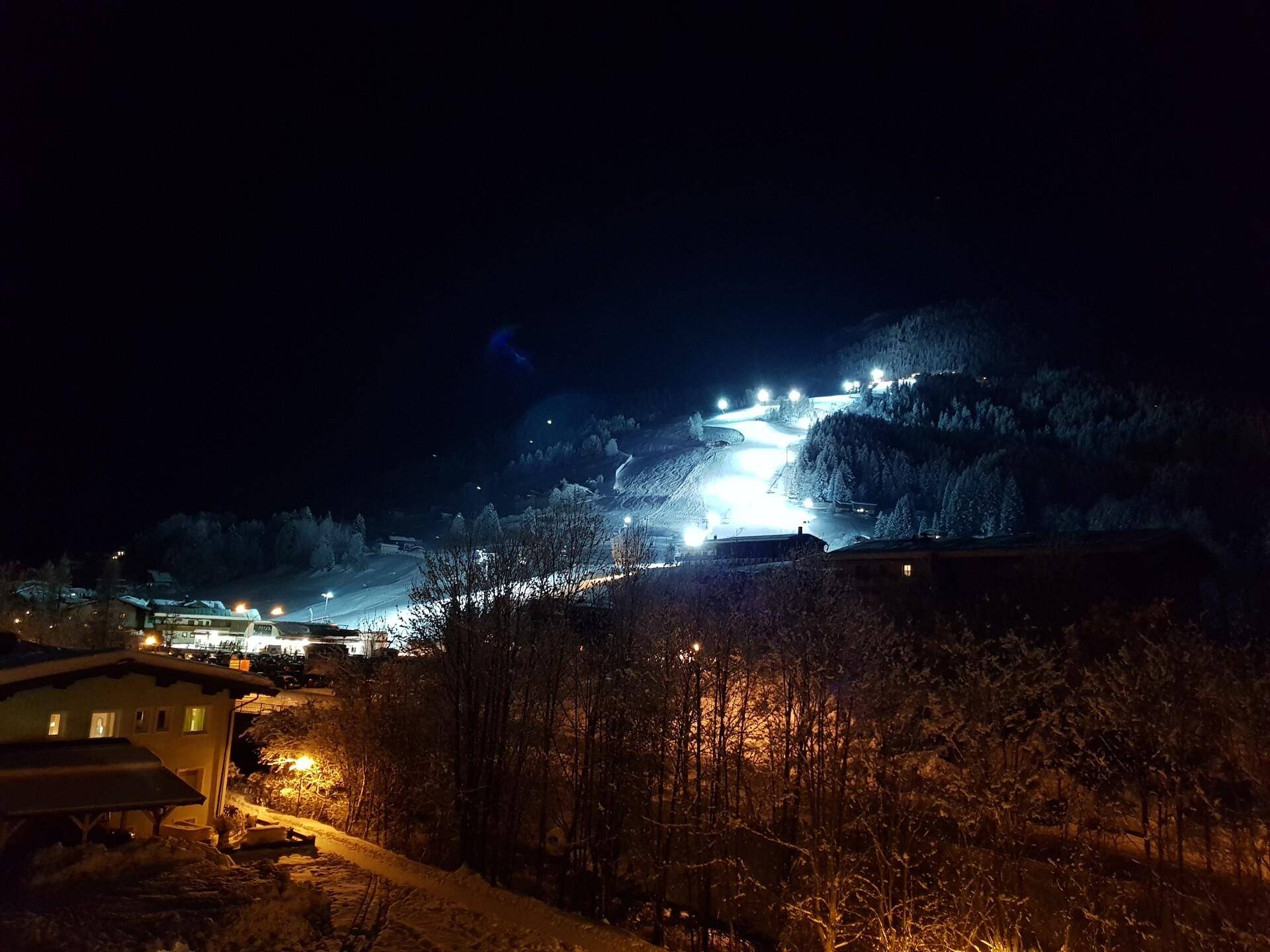 Beleuchtete Skipiste bei Nacht mit verschneiten Bergen und Häusern im Vordergrund.