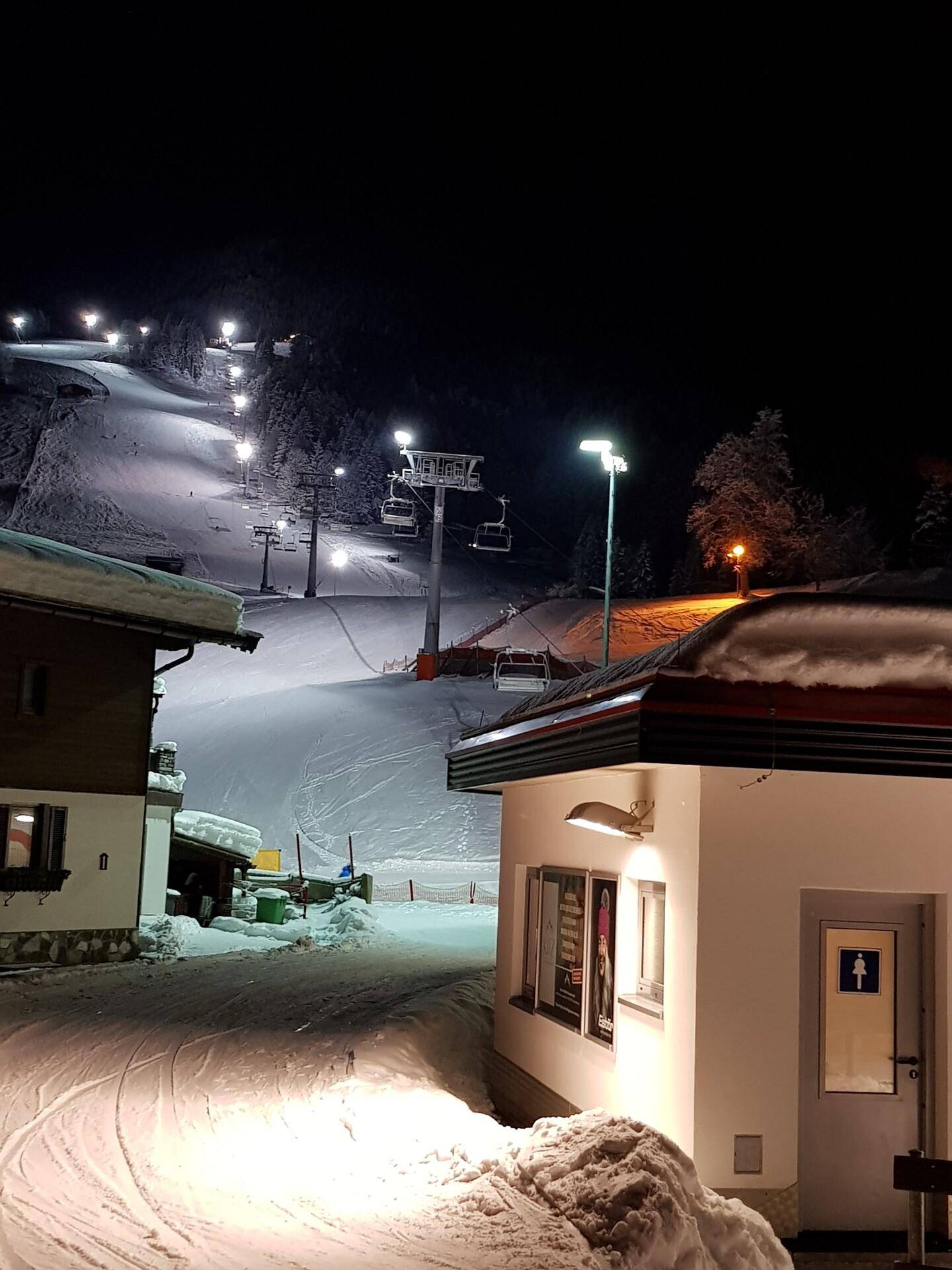 Beleuchteter Skilift und Piste bei Nacht mit verschneiter Umgebung und Gebäuden.