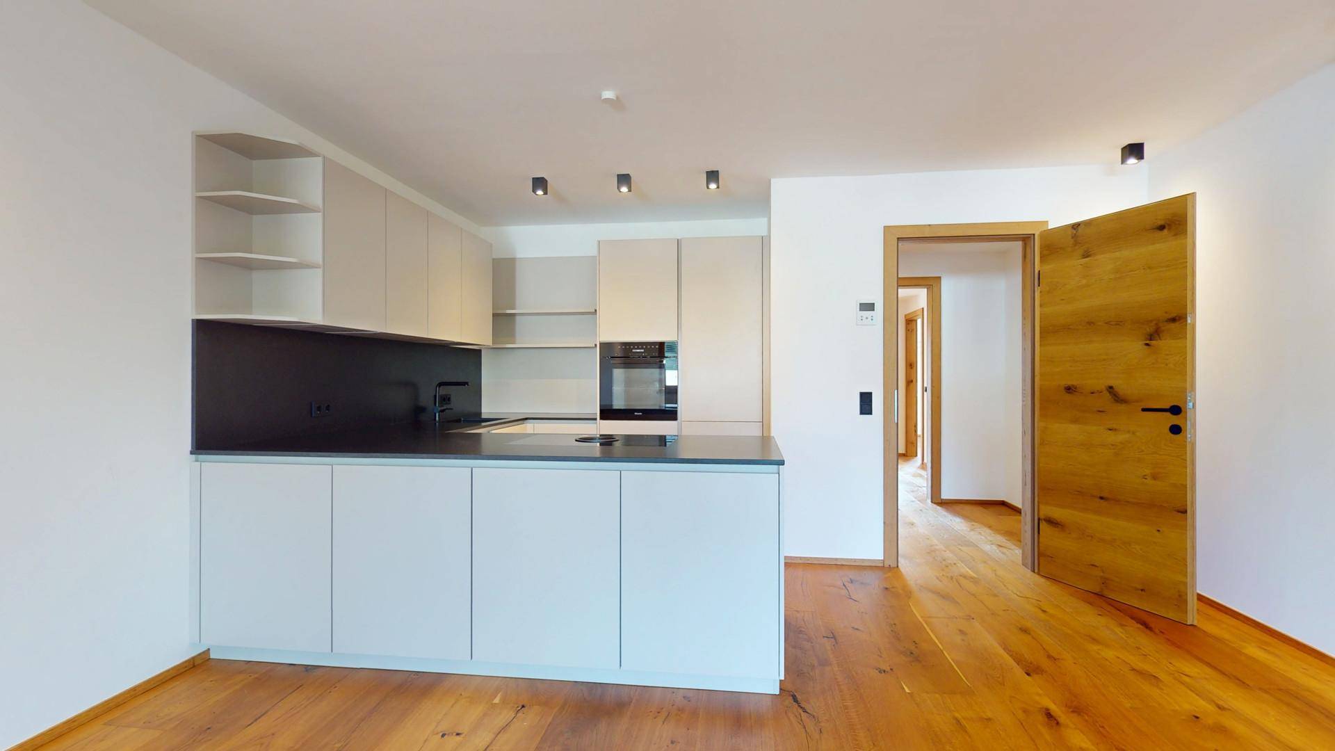 Moderne Einbauküche mit hellen Fronten, dunkler Arbeitsplatte und Holzboden in offenem Raum.