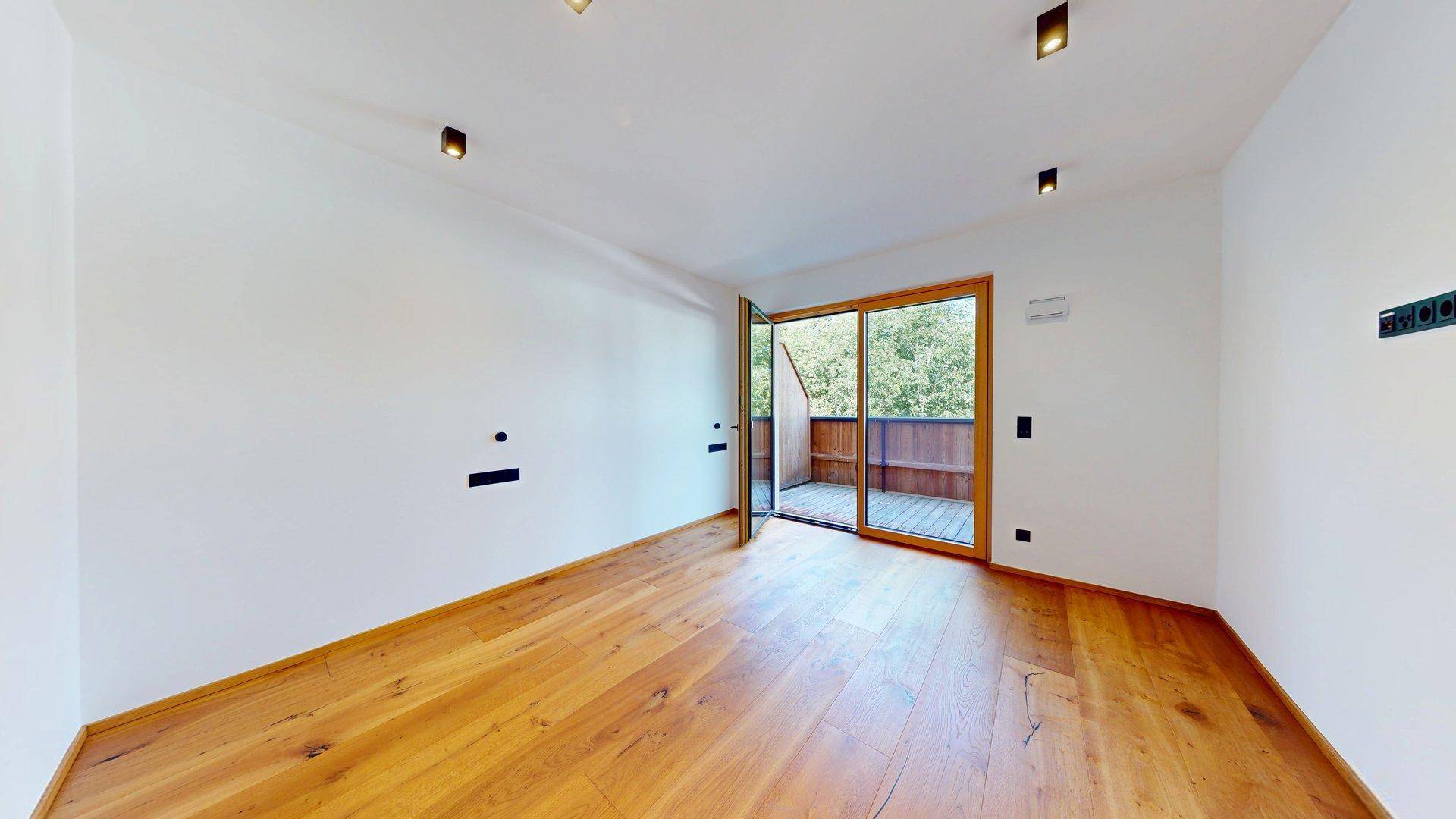 Geräumiges Zimmer mit hellem Holzboden und großem Fenster mit Blick ins Grüne.