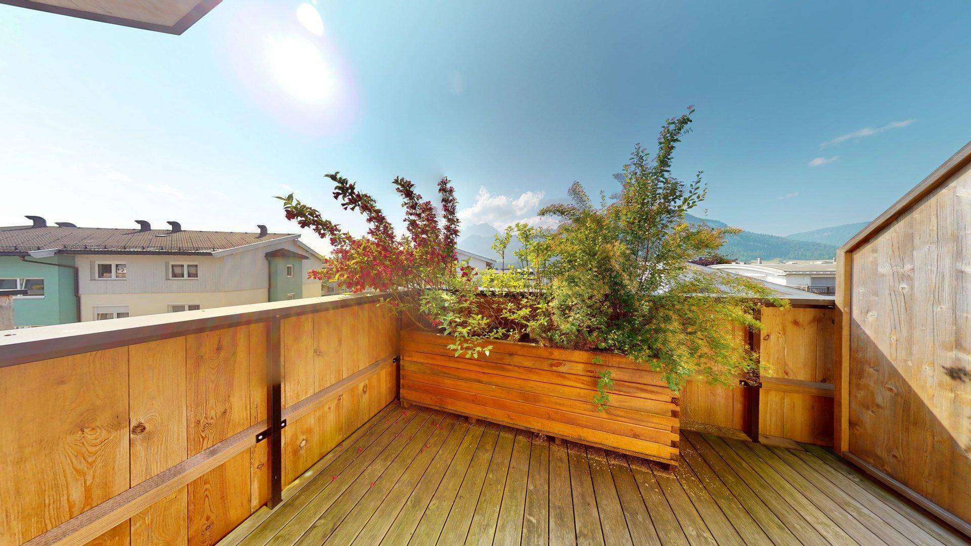 Sonniger Balkon mit Holzboden, Pflanzenkübeln und weitem Blick auf die Berge.
